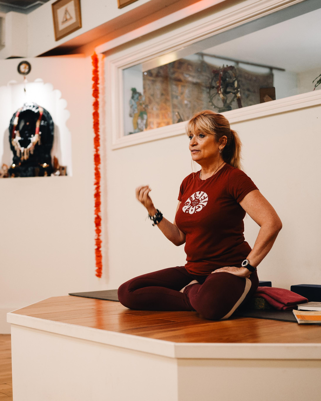 Jayne Orton in Iyengar Yoga Institute of Birmingham. A lady in a red t-shirt is sitting on the platform at the yoga studio. Yoga brand photoshoot.