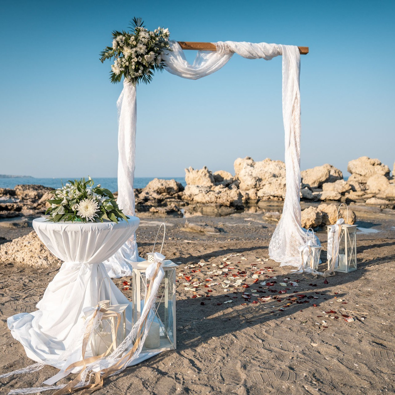 Romantische Hochzeitslocation am Strand von Faliraki-Rhodos: Dekorierter Traubogen mit weißen Blumen, Laternen und Blütenblättern auf dem Sand. Strandhochzeit am Meer mit Felsküste. Elegante Hochzeitsdekoration im Freien unter blauem Himmel.