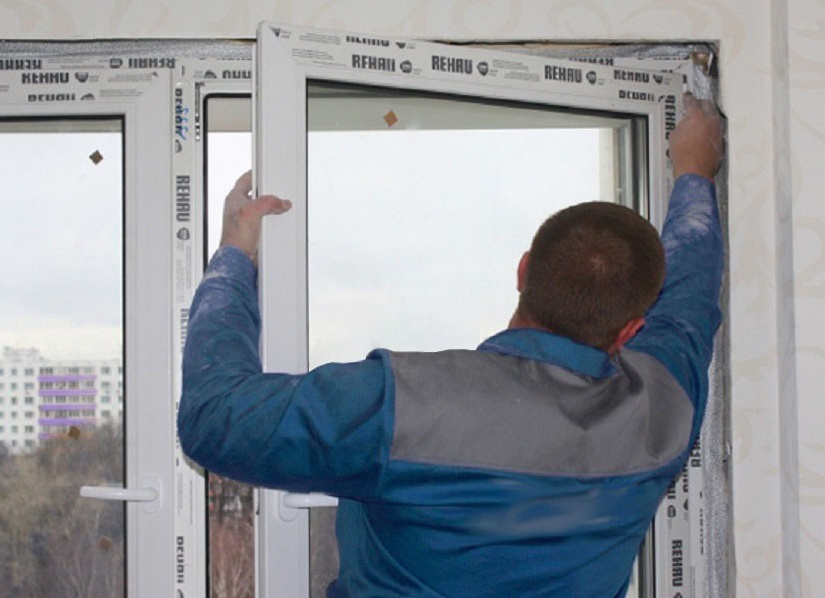 Монтажник в синей форменной куртке устанавливает пластиковое окно белого цвета фирмы Rehau.