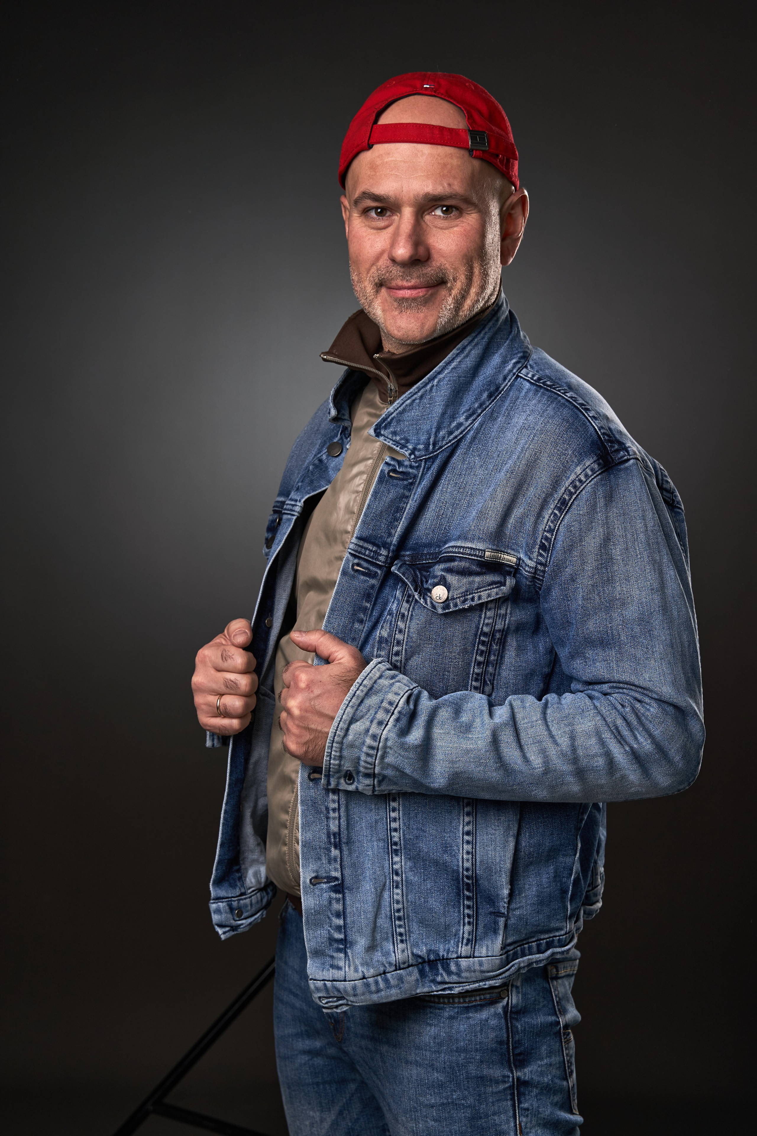 Man in a denim jacket and red baseball cap against a dark background