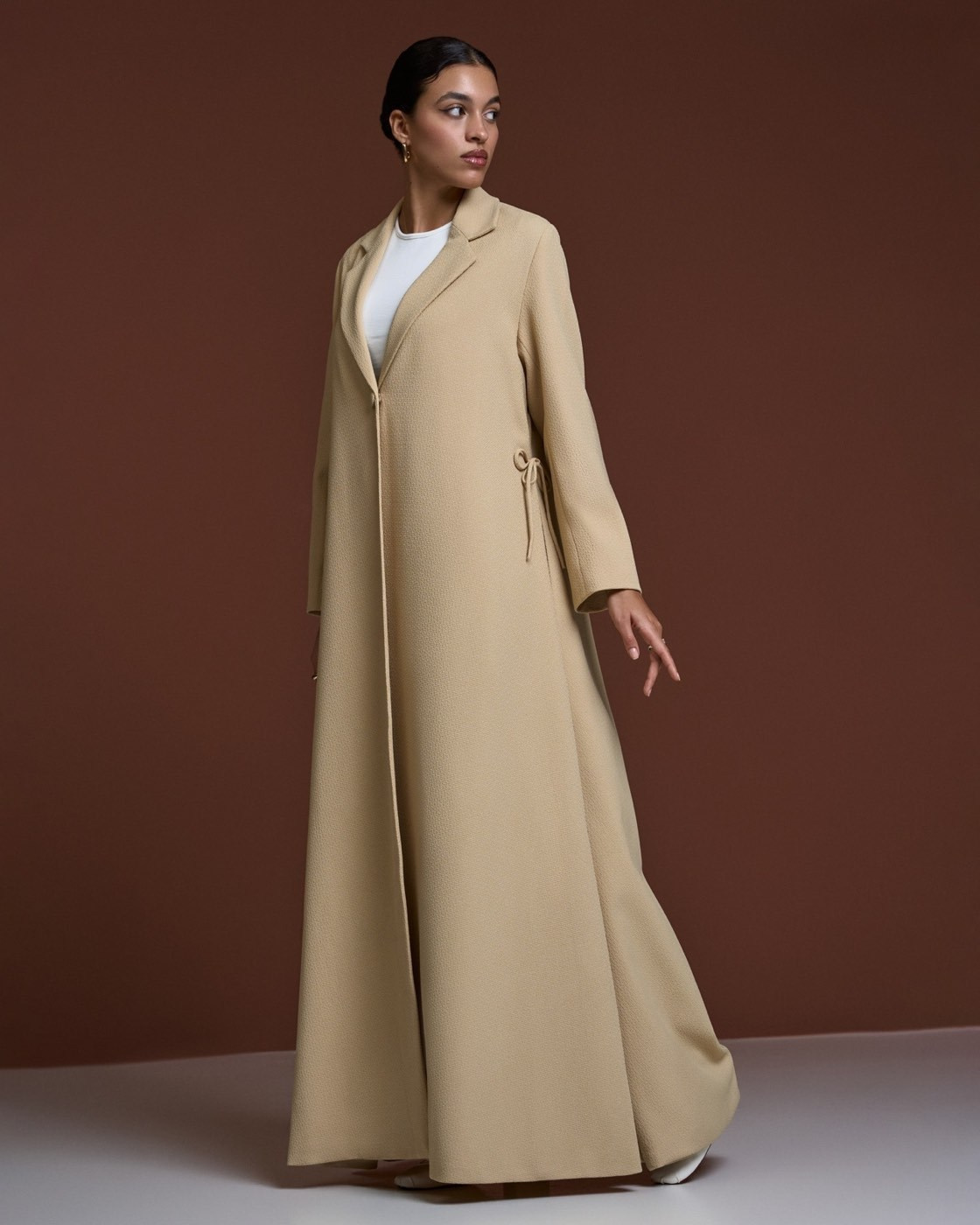 Model wearing a long beige coat over a white top against a brown studio background
