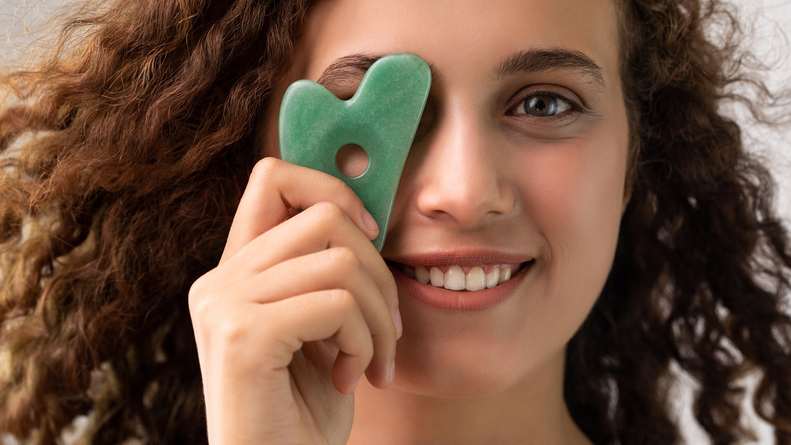 A smiling woman with curly hair holds a green gua sha tool over one eye, promoting facial massage and skincare.