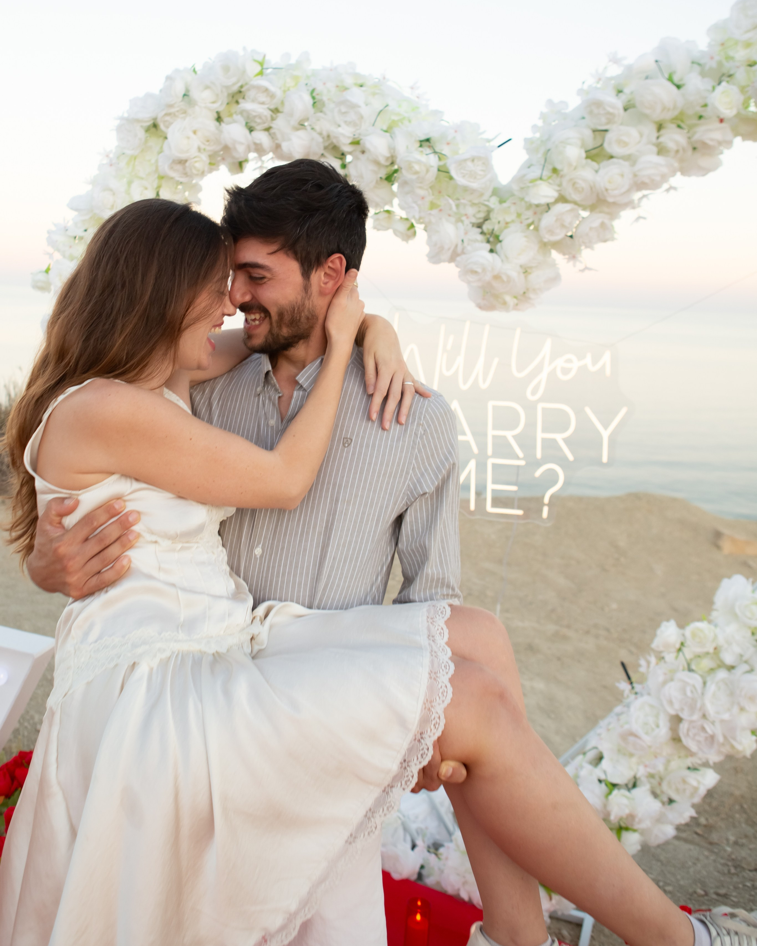 Happy couple celebrating a romantic marriage proposal beside a red heart decoration on the beach in Spain, Benidorm