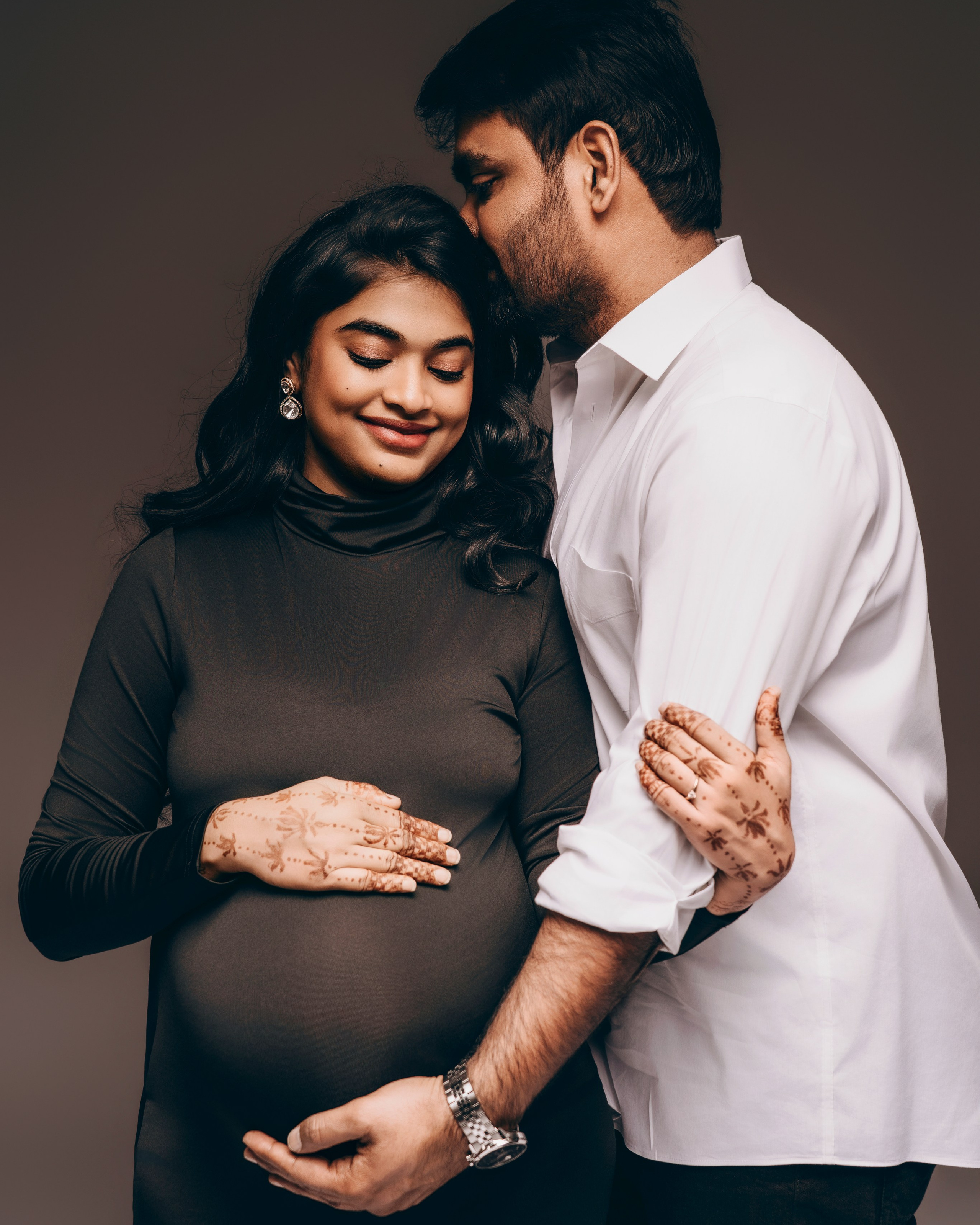 Intimate maternity couple portrait, husband kissing pregnant wife's forehead, henna hands on belly.