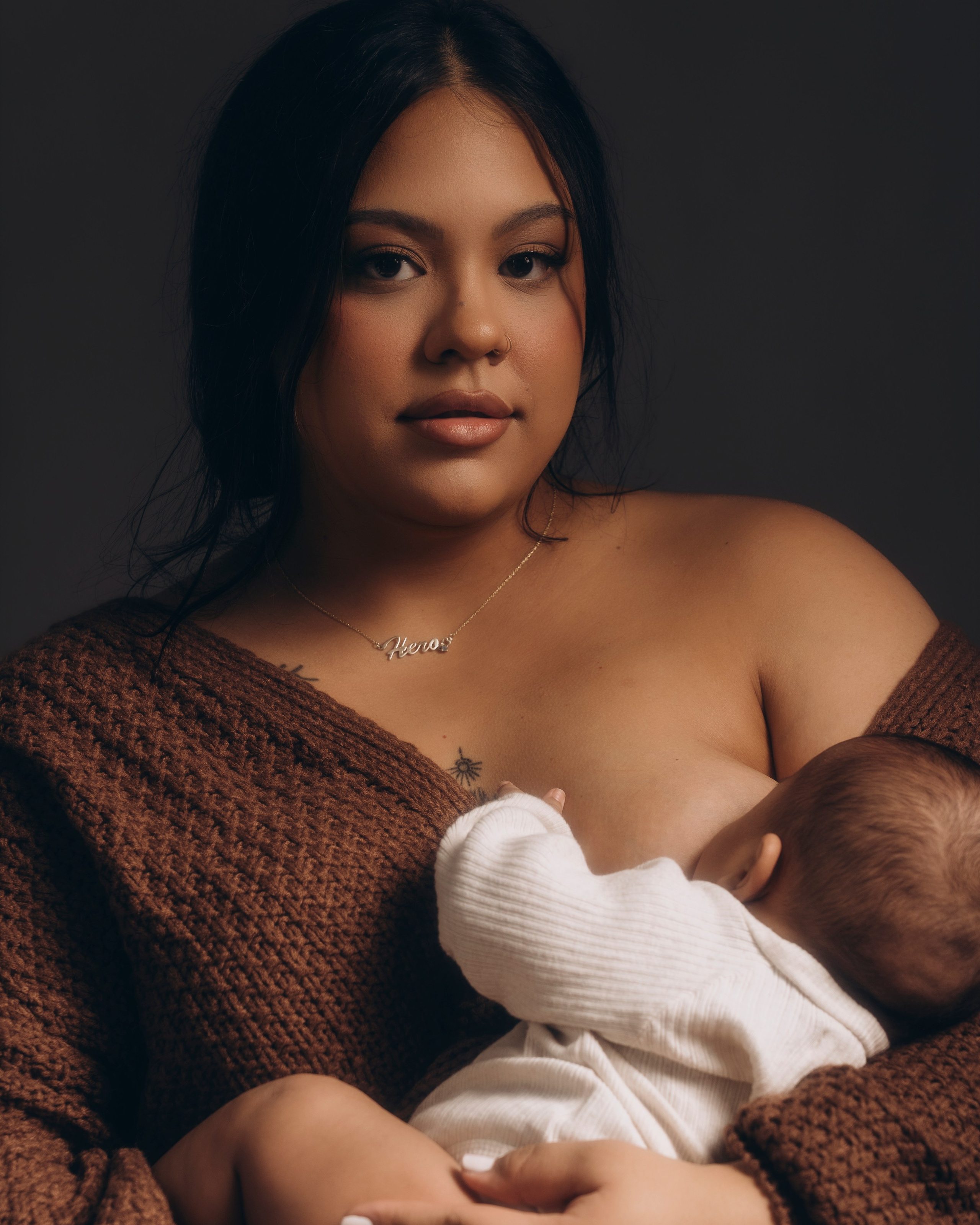 Editorial breastfeeding portrait, mother nursing baby in brown knit cardigan, dark studio mood.