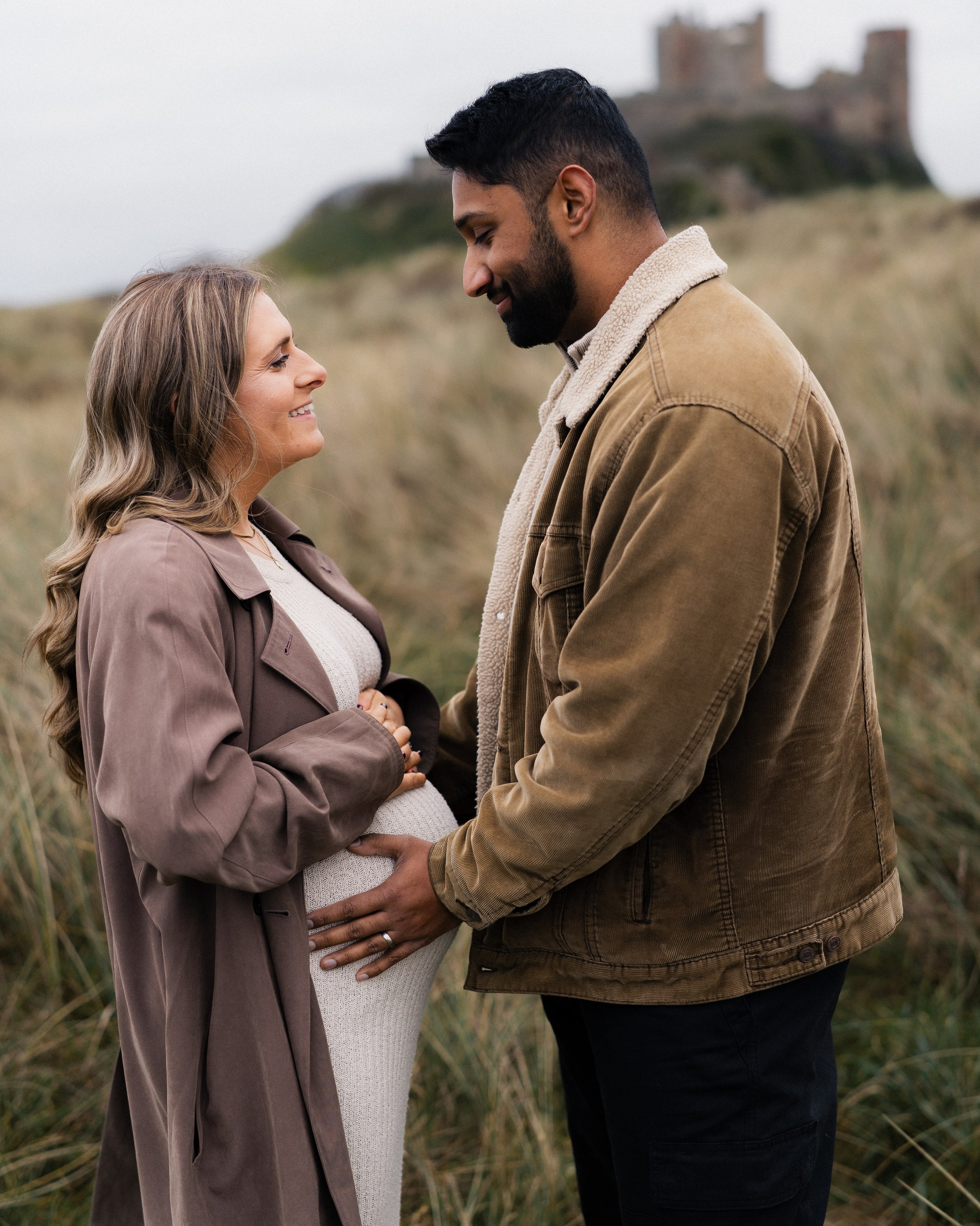 Editorial maternity photography in Bamburgh beach, in Northumberland. Shot with natural light photograph of a couple embracing in tall grass.