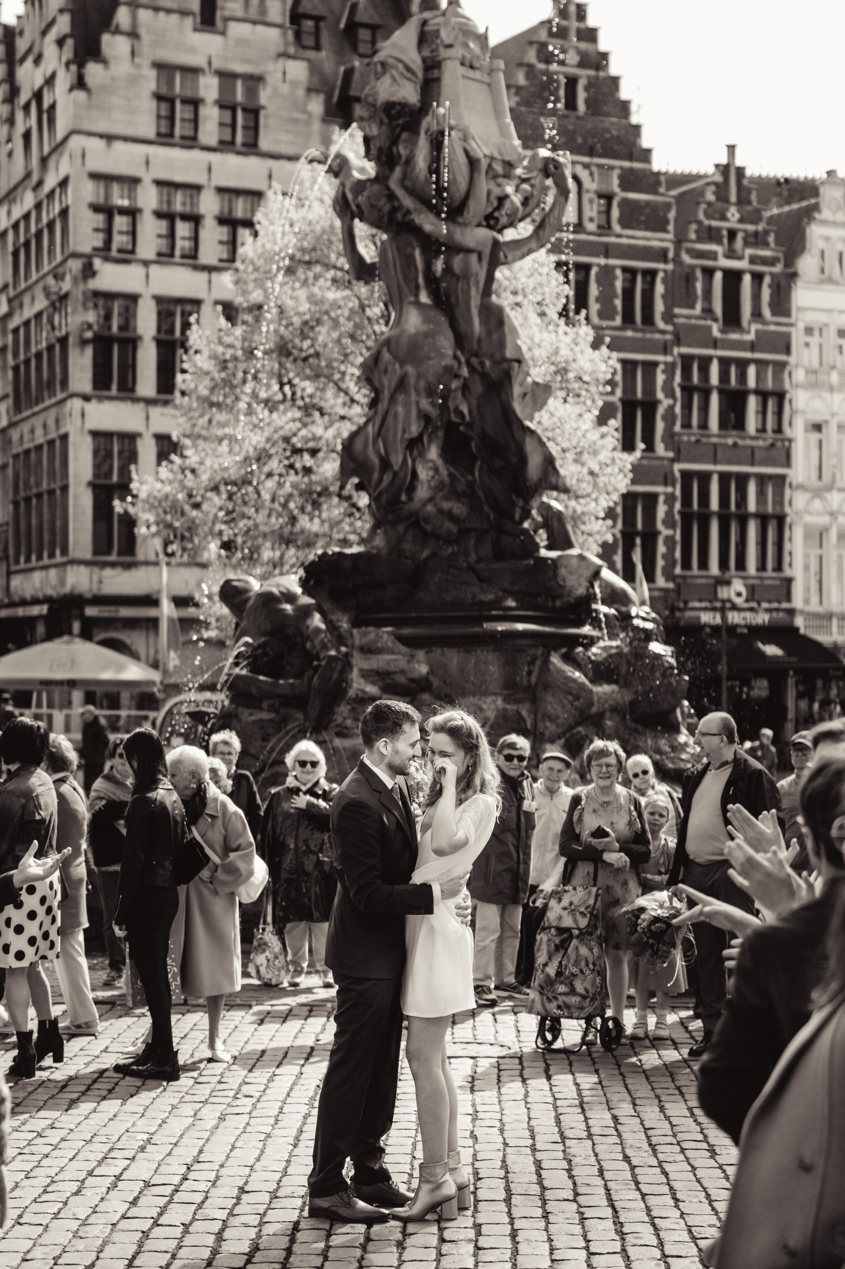 Het bruidspaar staat samen tussen familieleden op de Grote Markt in Antwerpen, tijdens een ontroerend moment waarop de bruid zichtbaar ontroerd is.