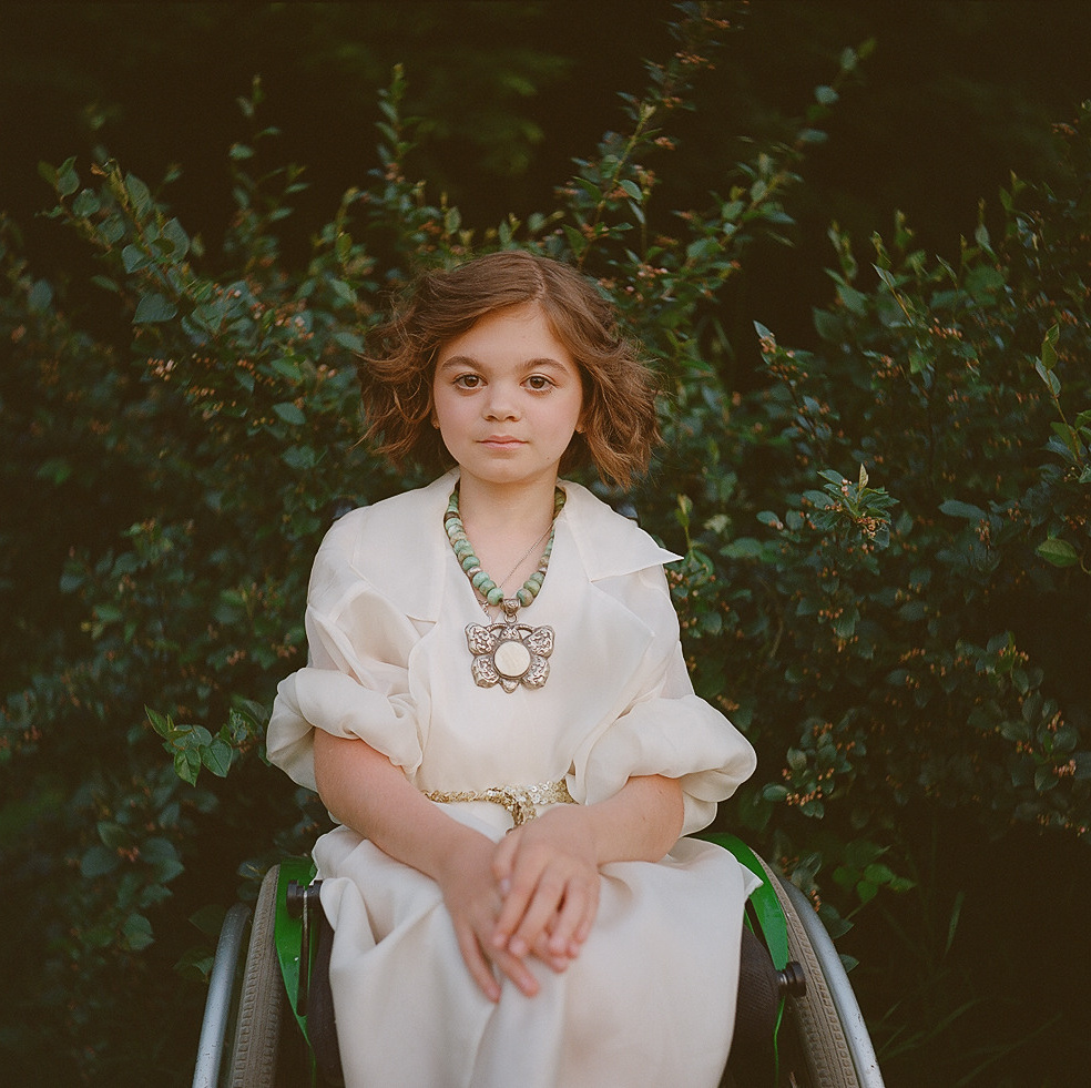 Girl is sitting in a wheel chair in a beautiful outfit with a bush on a background, from wheeling heart project