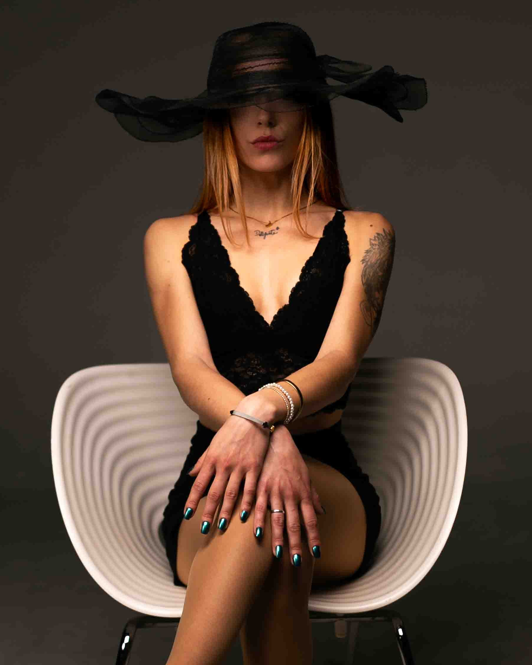 Modelo vestida de negro con pamela negra y sentada en un sillón blanco