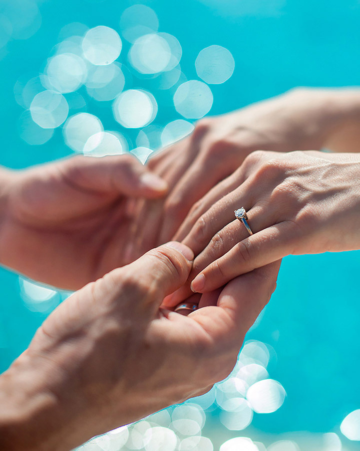Фотография свадебных колец на руках пары на фоне яркого и бирюзового океана.