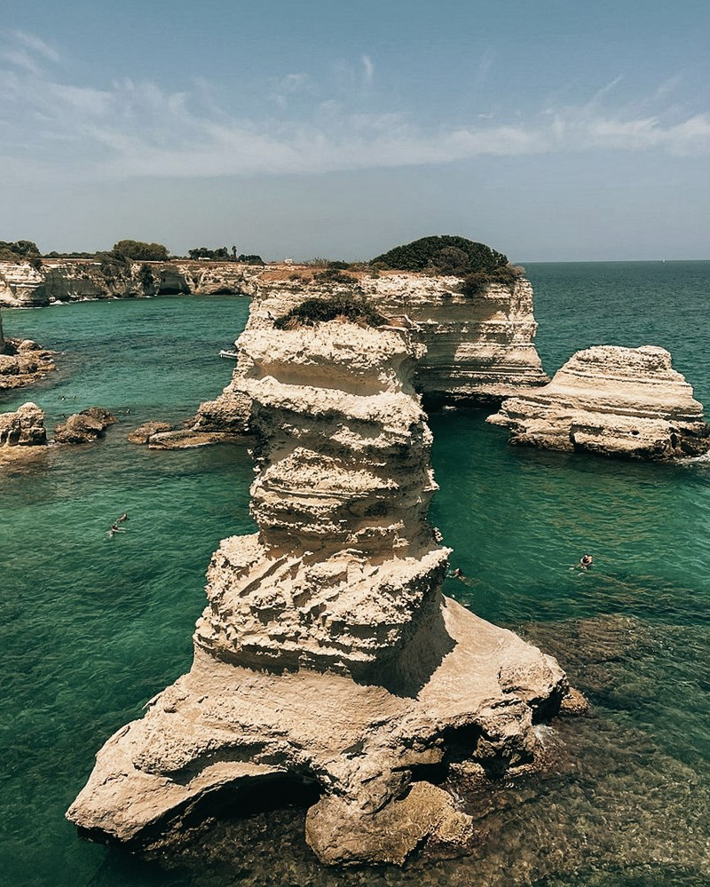 The dramatic cliffs of Torre Sant’Andrea rise above turquoise water. Swimmers relax at the base of the rocks under the blazing summer sun.