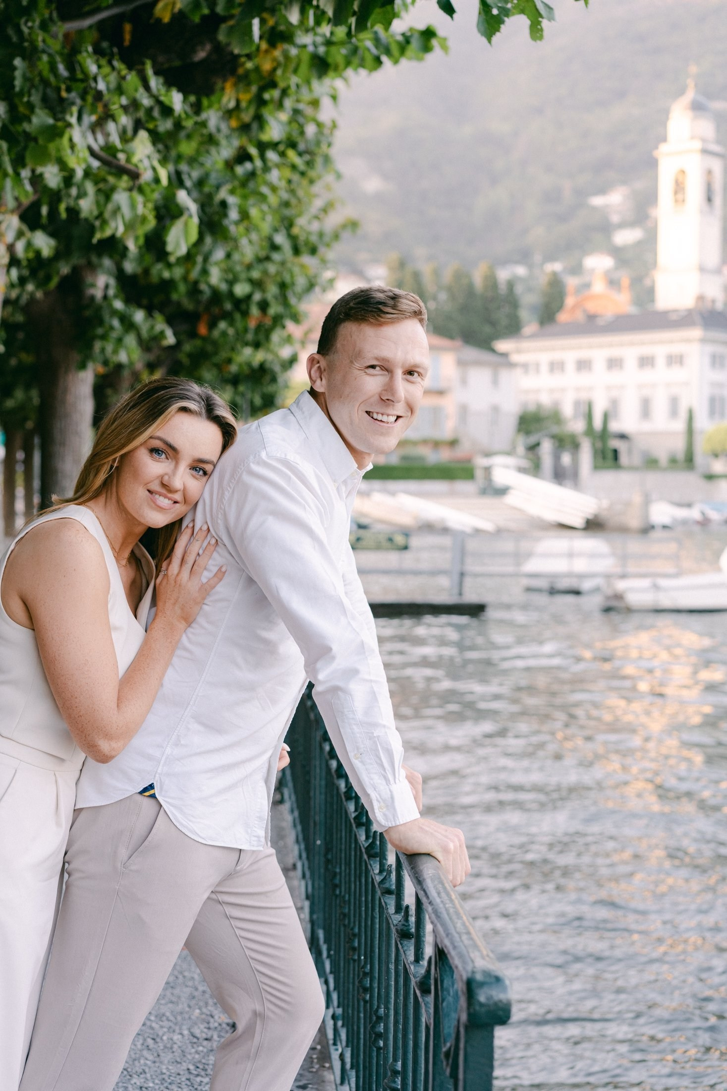 Couple posing by the Lake Como waterfront with a historic village and church tower in the background
