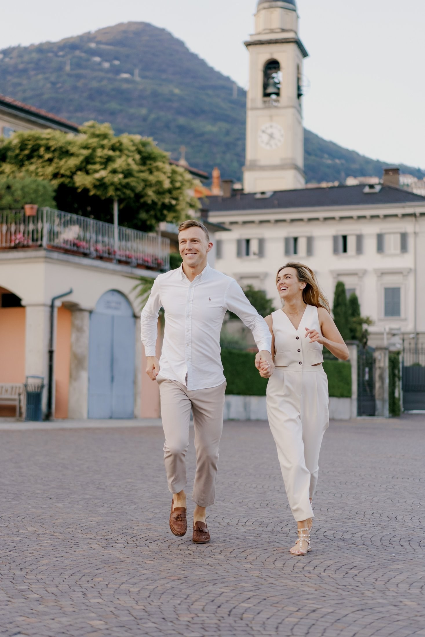 Smiling couple running hand in hand through a Lake Como village square with a church tower and mountains behind them