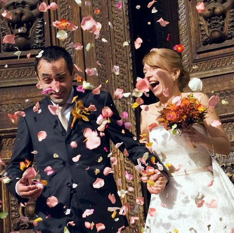 Red-haired bride holding her flowing veil aloft smiles seductively to her bouquet holding groom as he leans back against a railing in appreciation at the Vrtba Garden in Prague. 