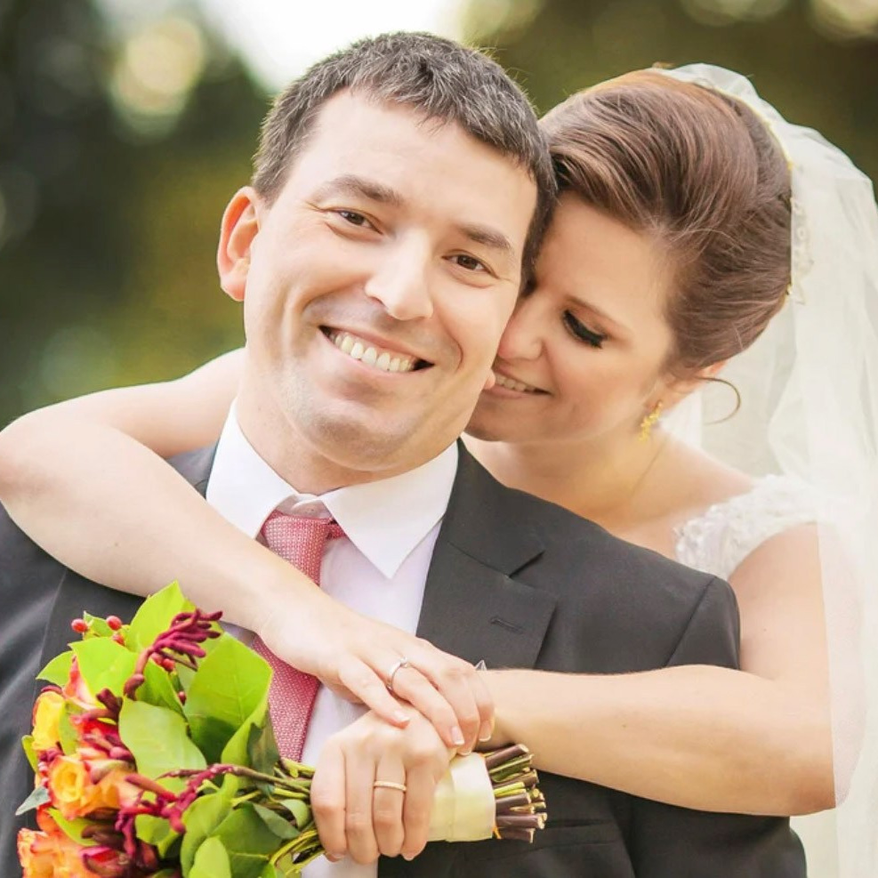Smiling, radiant Mexican bride hugs her groom from behing while holding her bouquet.