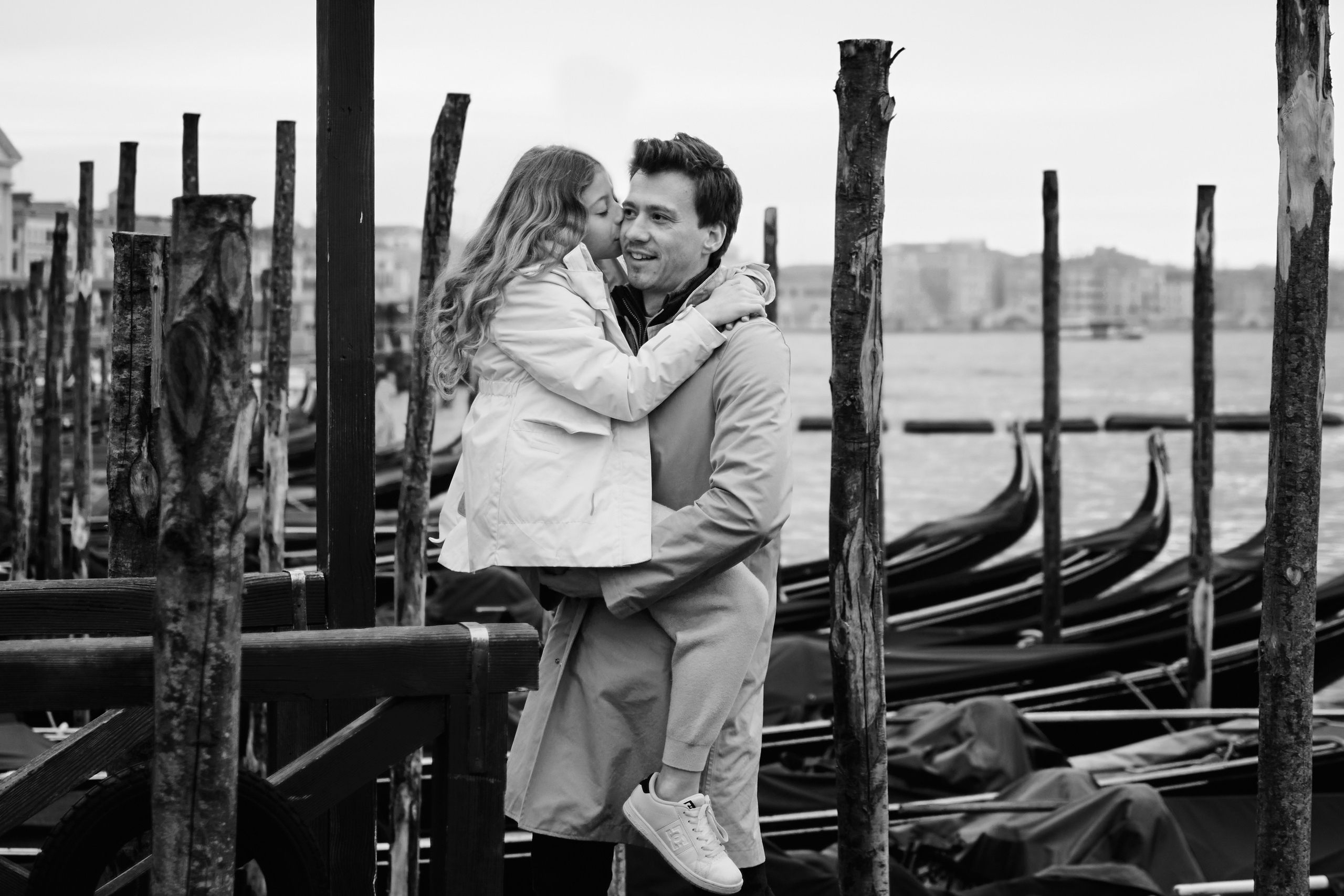 A happy family of four stands in front of the iconic St. Mark's Square, with the beautiful Venetian architecture and gondolas in the background. Against the backdrop of San Marco Square in Italy, a dad and his child share a moment of joy and wonder in Venice.