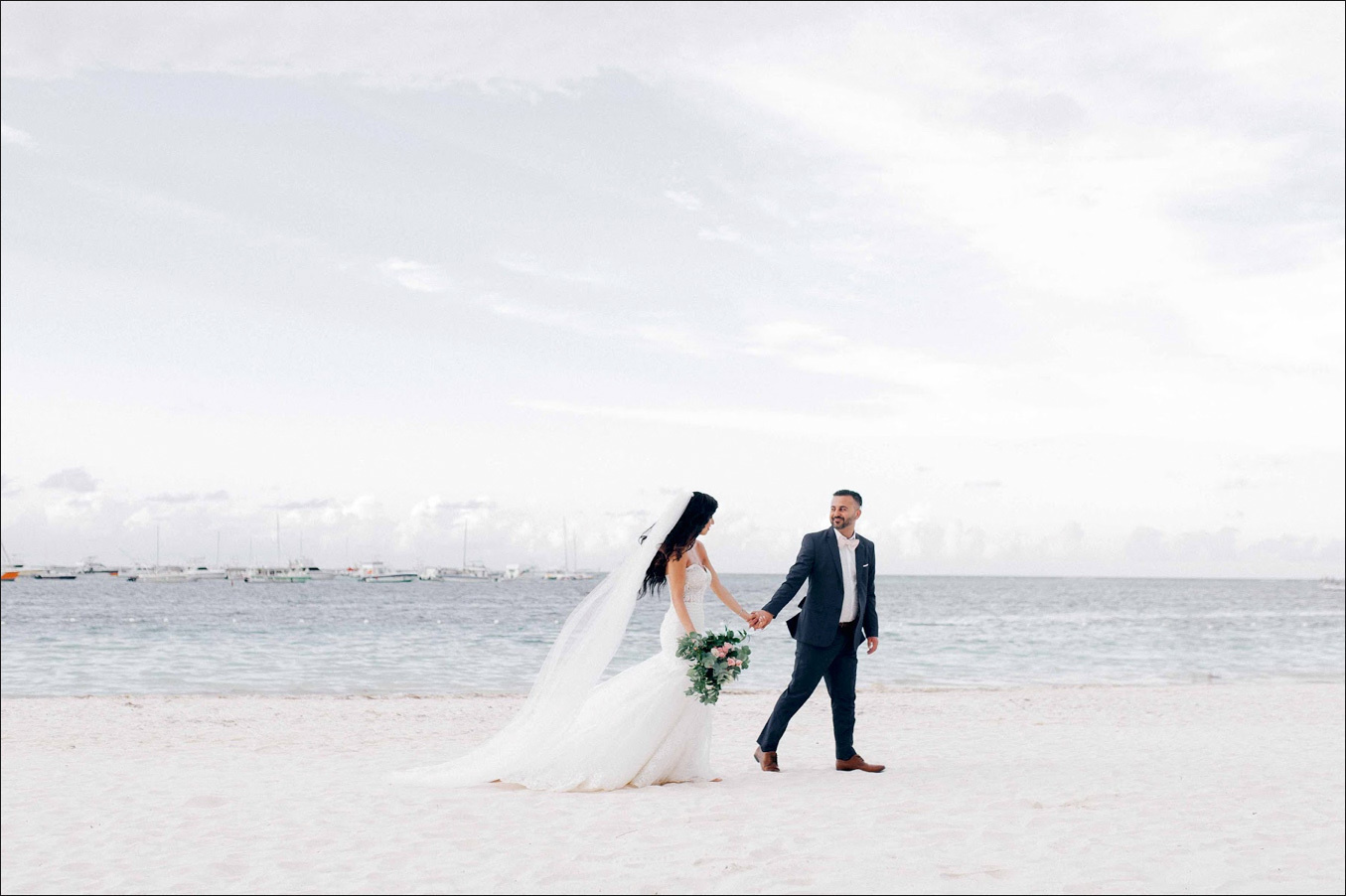 Невеста ведет невесту за руку по пляжу, у невесты классическое свадебное платье и фата развивается на ветру. 