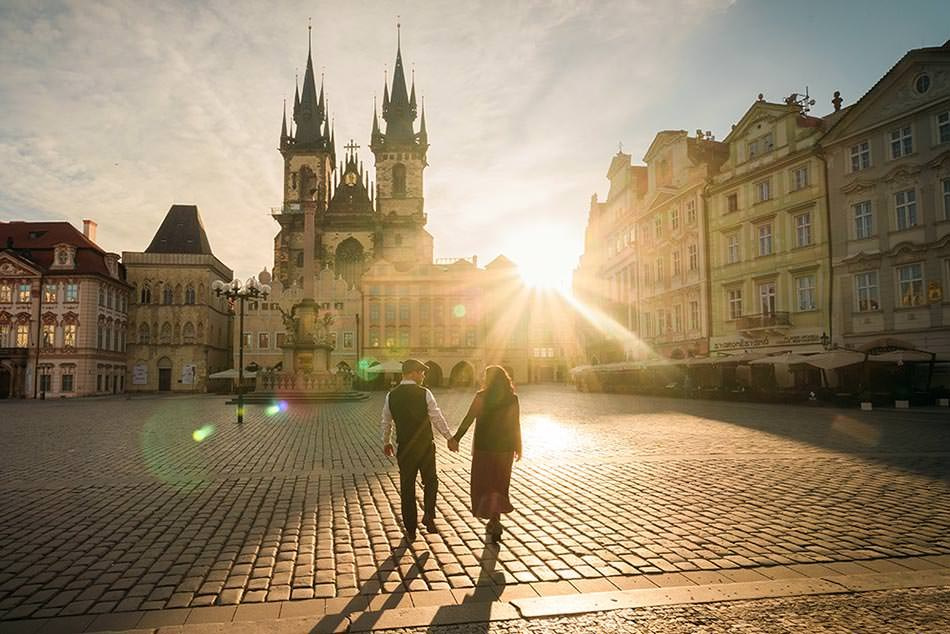 An American couple walks towards the early morning sunshine as it breaks above the Old Town Square in Prague during their engagement session.