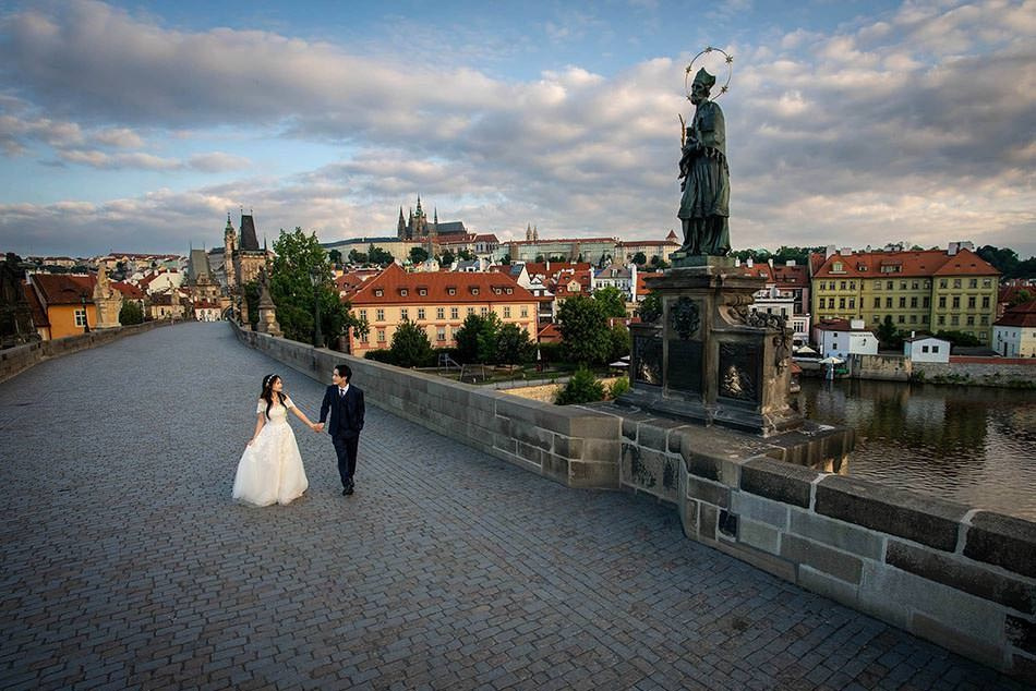 A young Korean bride is escorted by her groom atop the deserted Charles Bridge during their sunrise pre-wedding in Prague. 