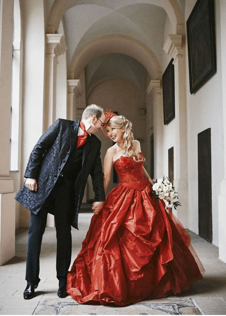 Stylish English groom touching foreheads with blonde bride in red dress under historic Prague church arches.