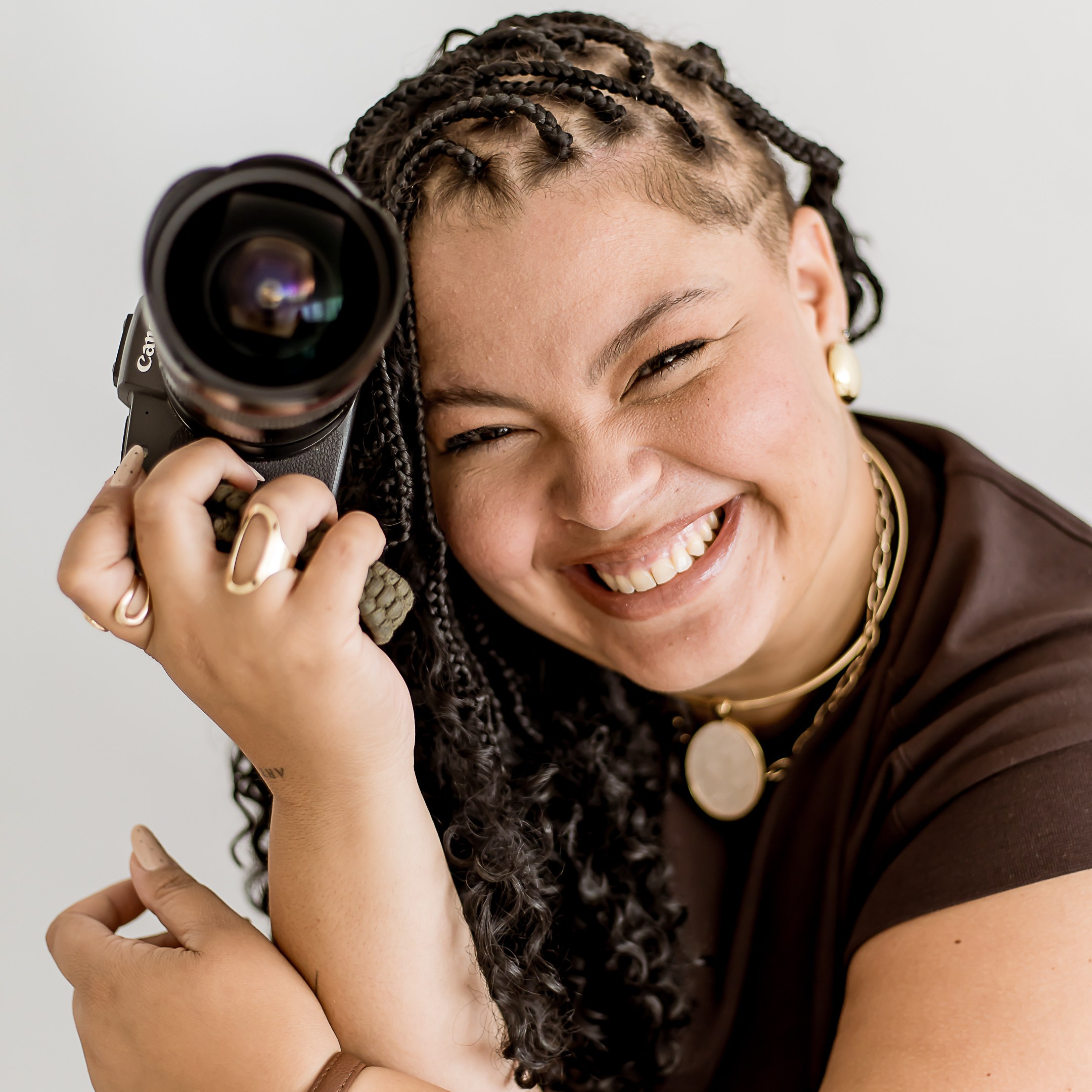 fotografa parda de tranças com camisa marrom sorrindo com camera na mão