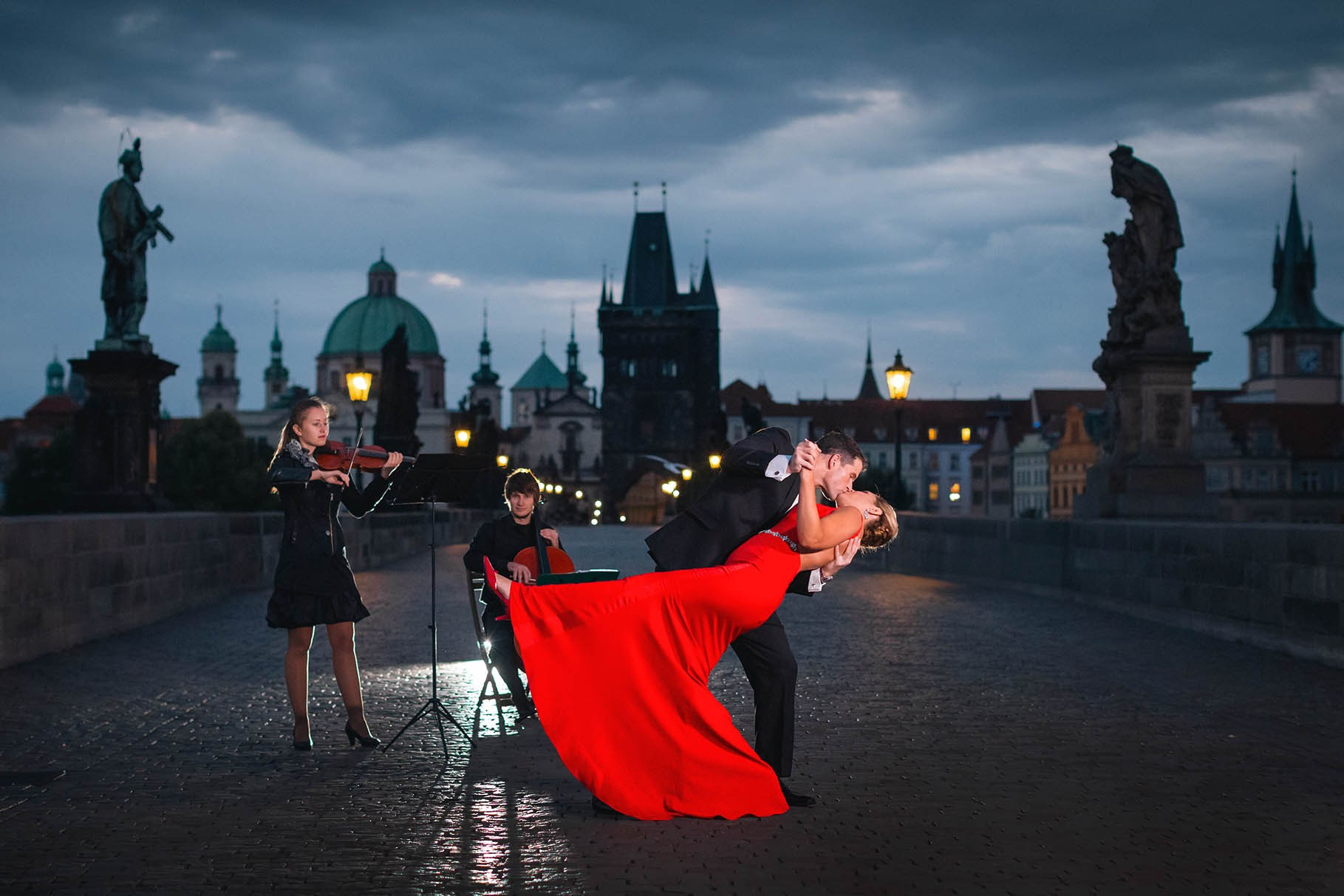 As a cellist and a violinist play, a woman in a red dress is romantically dipped for a kiss atop the Charles Bridge moments after she agreed to marry him in Prague.