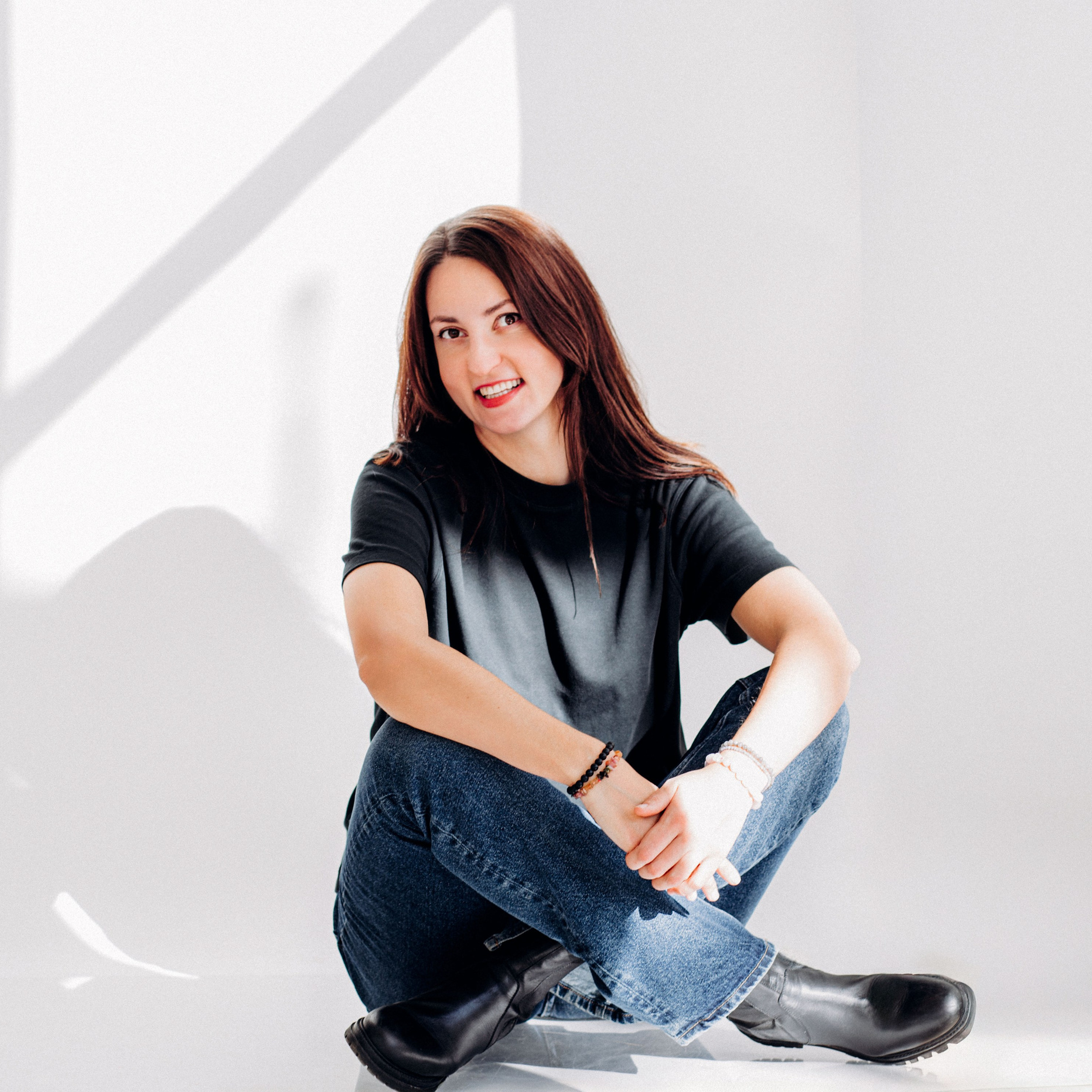Retrato con actitud de una mujer sentada de forma casual en el suelo de un estudio iluminado por el sol, vestida con camiseta negra, vaqueros y botas negras — una imagen moderna y relajada capturada en Valencia, España.
