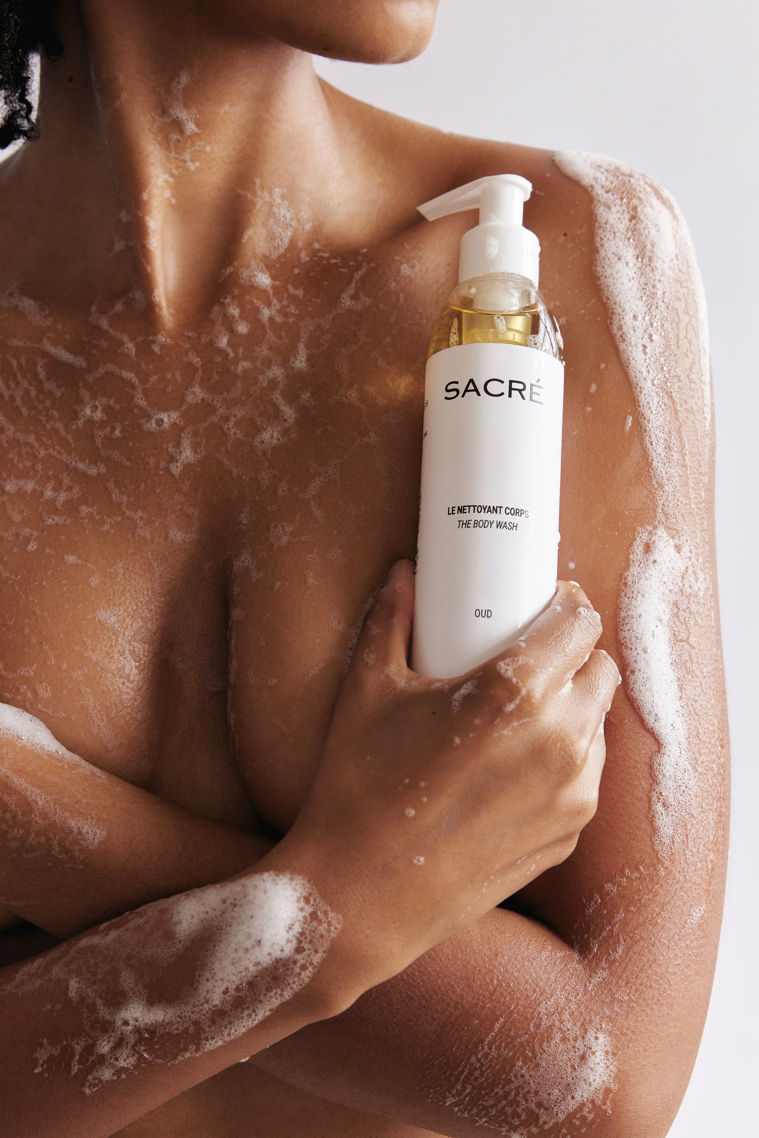 Model holding Sacré body wash while covered in soft foam, showcasing luxury skincare, natural beauty, and a refreshing self-care routine.