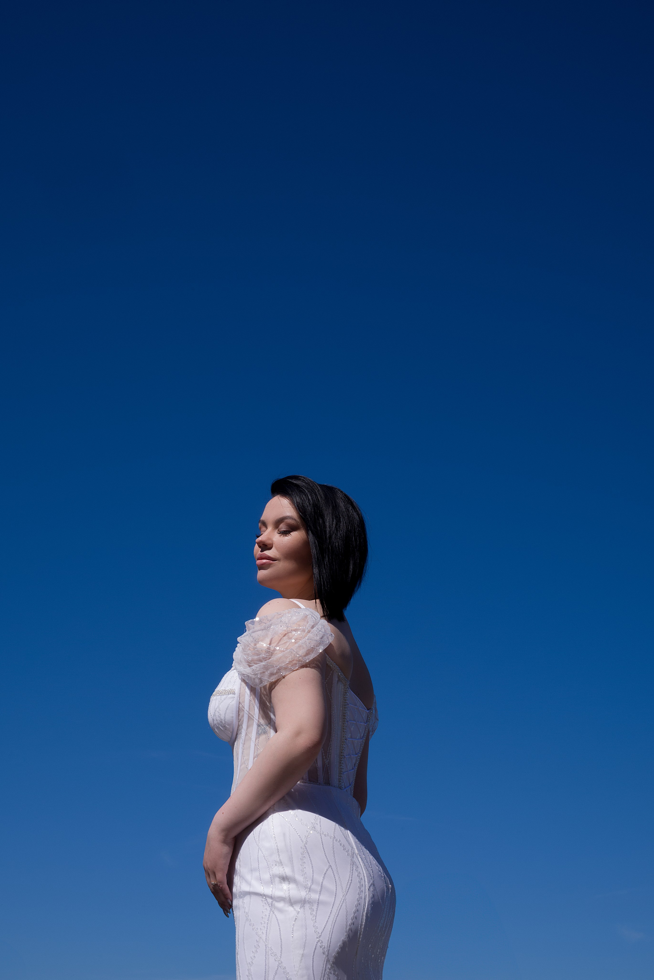 An elegant bride in a white wedding dress stands against a deep blue sky, her eyes closed and expression serene. The clean lines and minimalist composition create a cinematic and editorial feel.