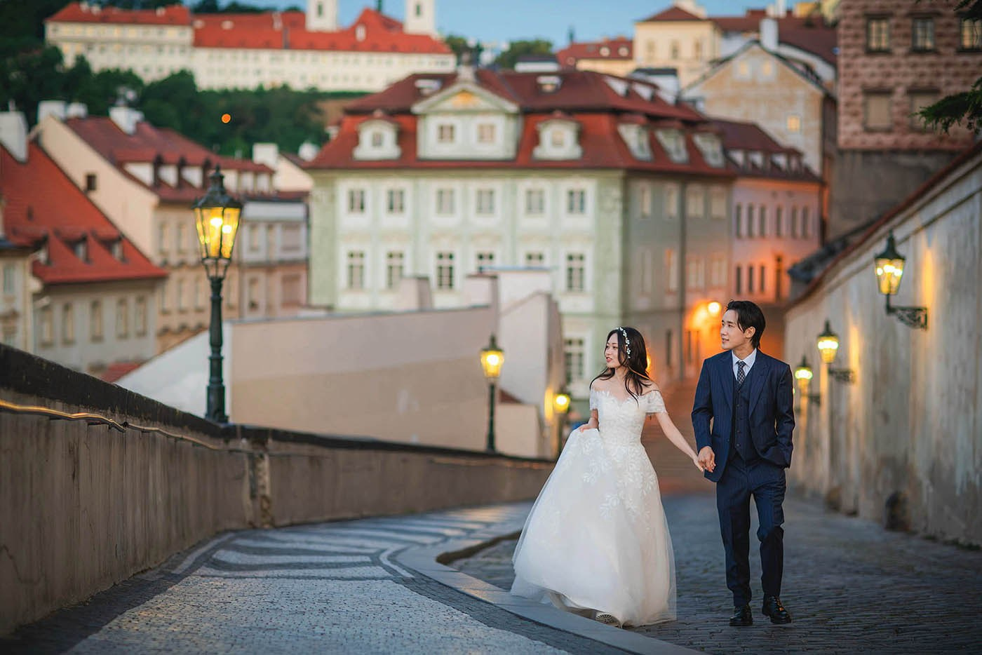 Newlyweds from Korea walk hand in hand through the gas lamp-lit landscape of Prague Castle before dawn during their pre-wedding