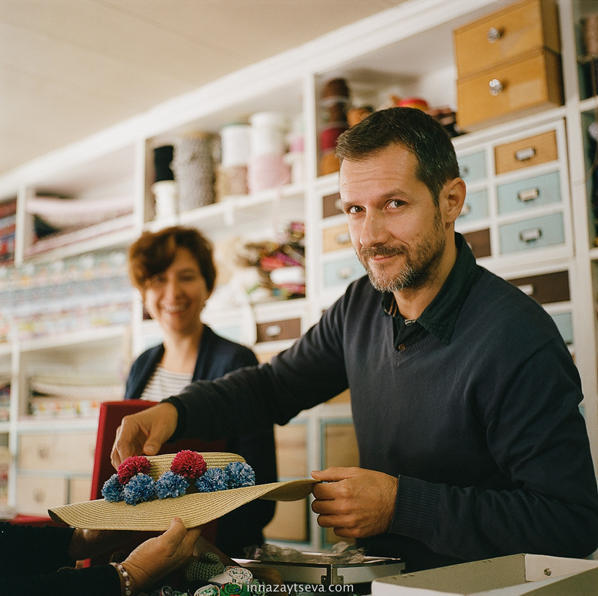 Mann repariert den Hut in seinem Werkstattatelier voller Schränke mit Material und seine Kollegin im Hintergrund