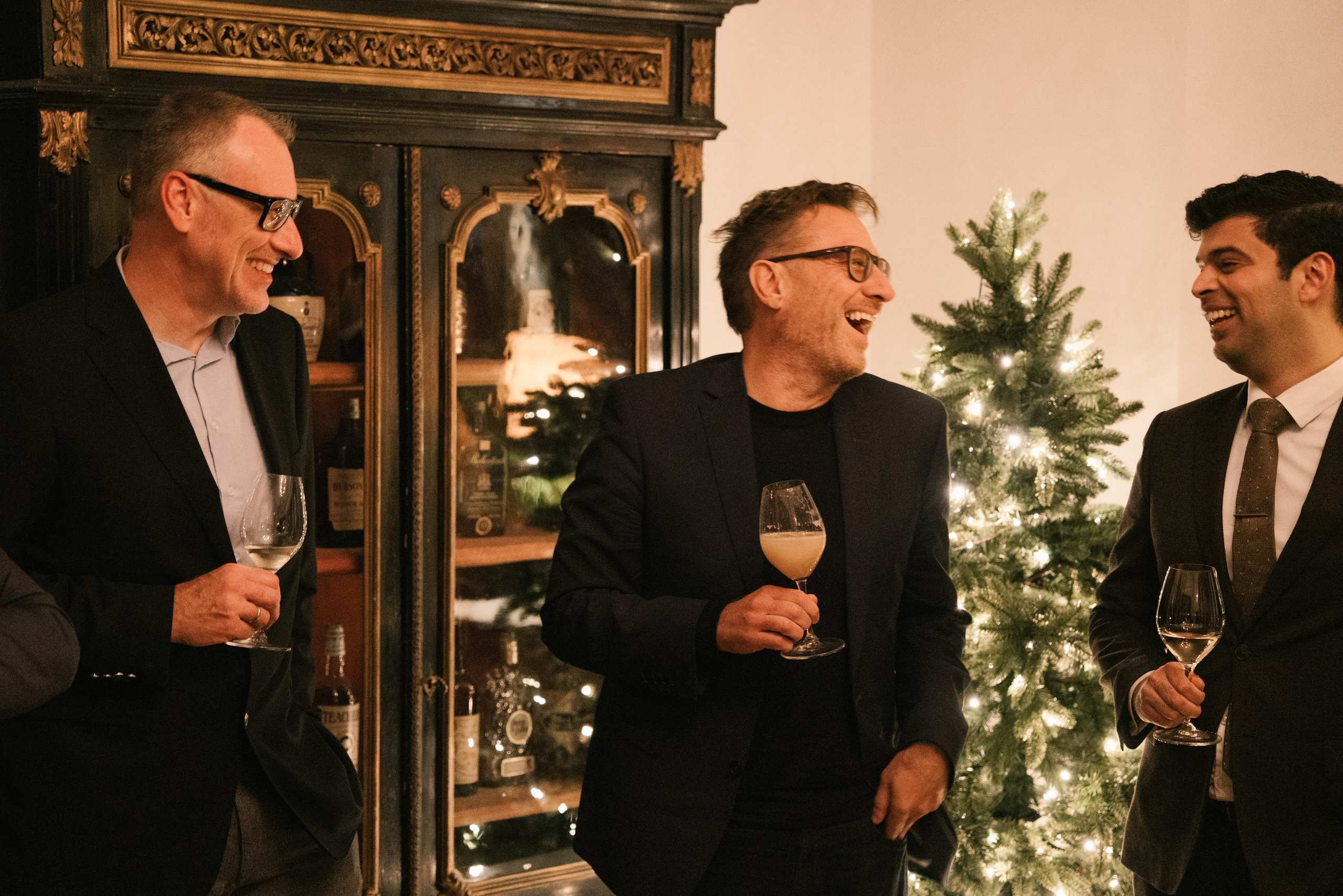 Men in suits laughing at an evening Christmas party near a lit Christmas tree