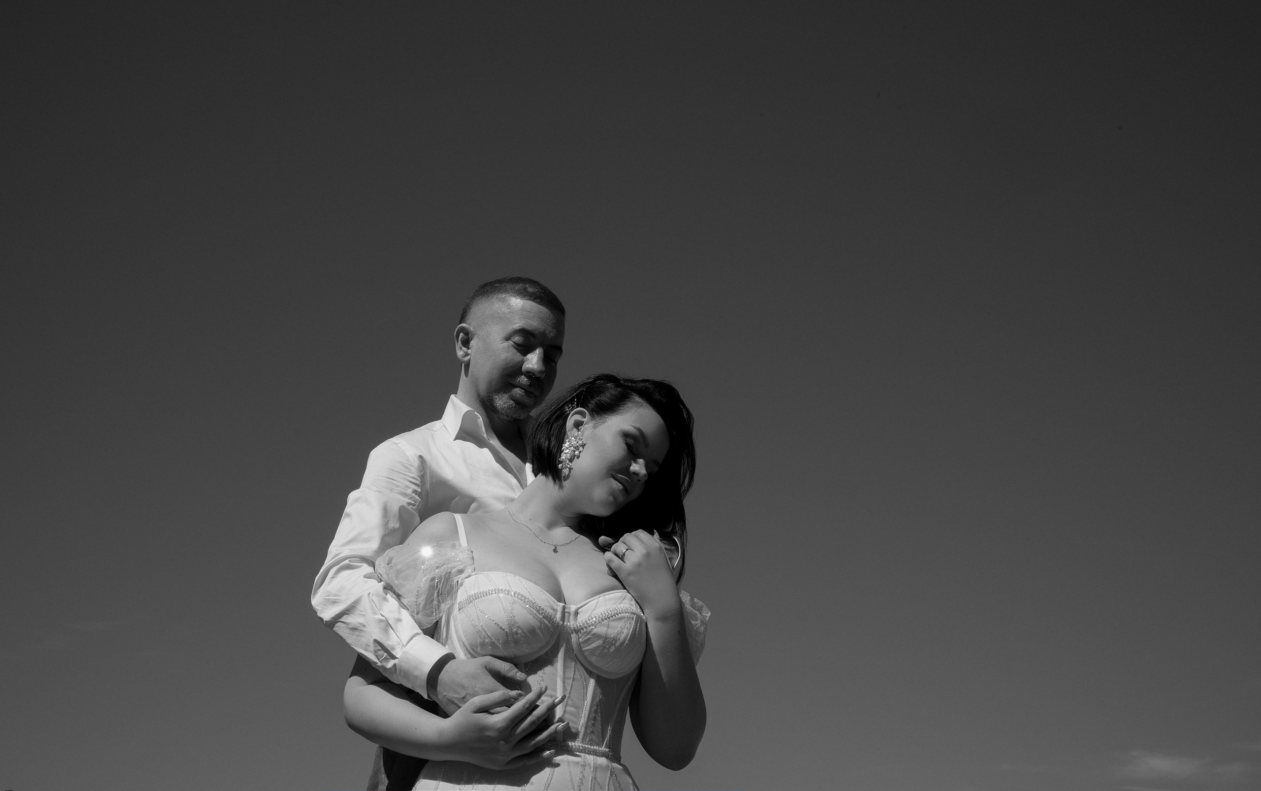 A romantic black and white portrait of a couple: the groom gently embraces the bride from behind as she leans into him with a soft, dreamy smile. The image captures intimacy and genuine connection.
