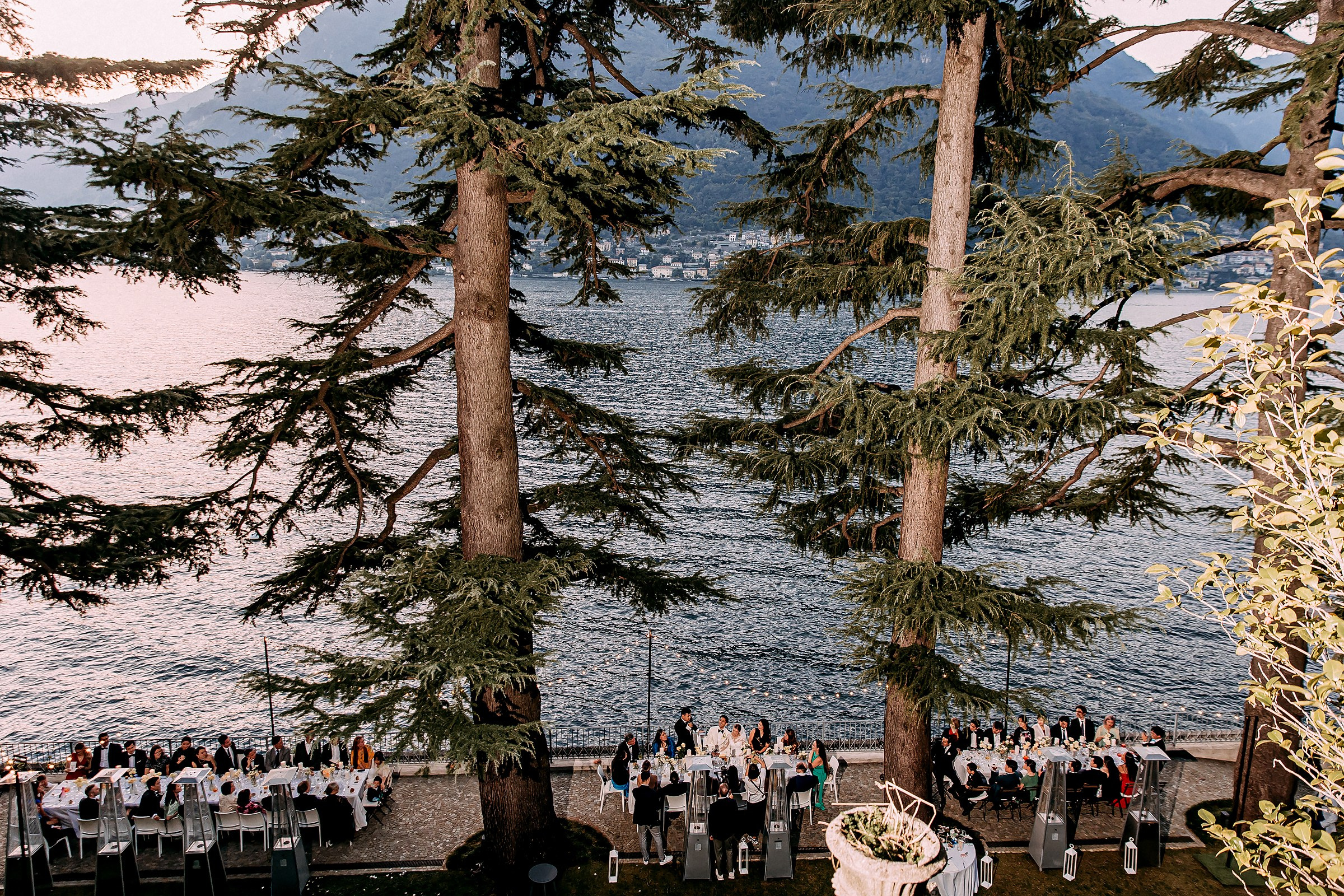 Scenic outdoor dining setup with string lights, surrounded by tall trees and overlooking a serene lake Como. 