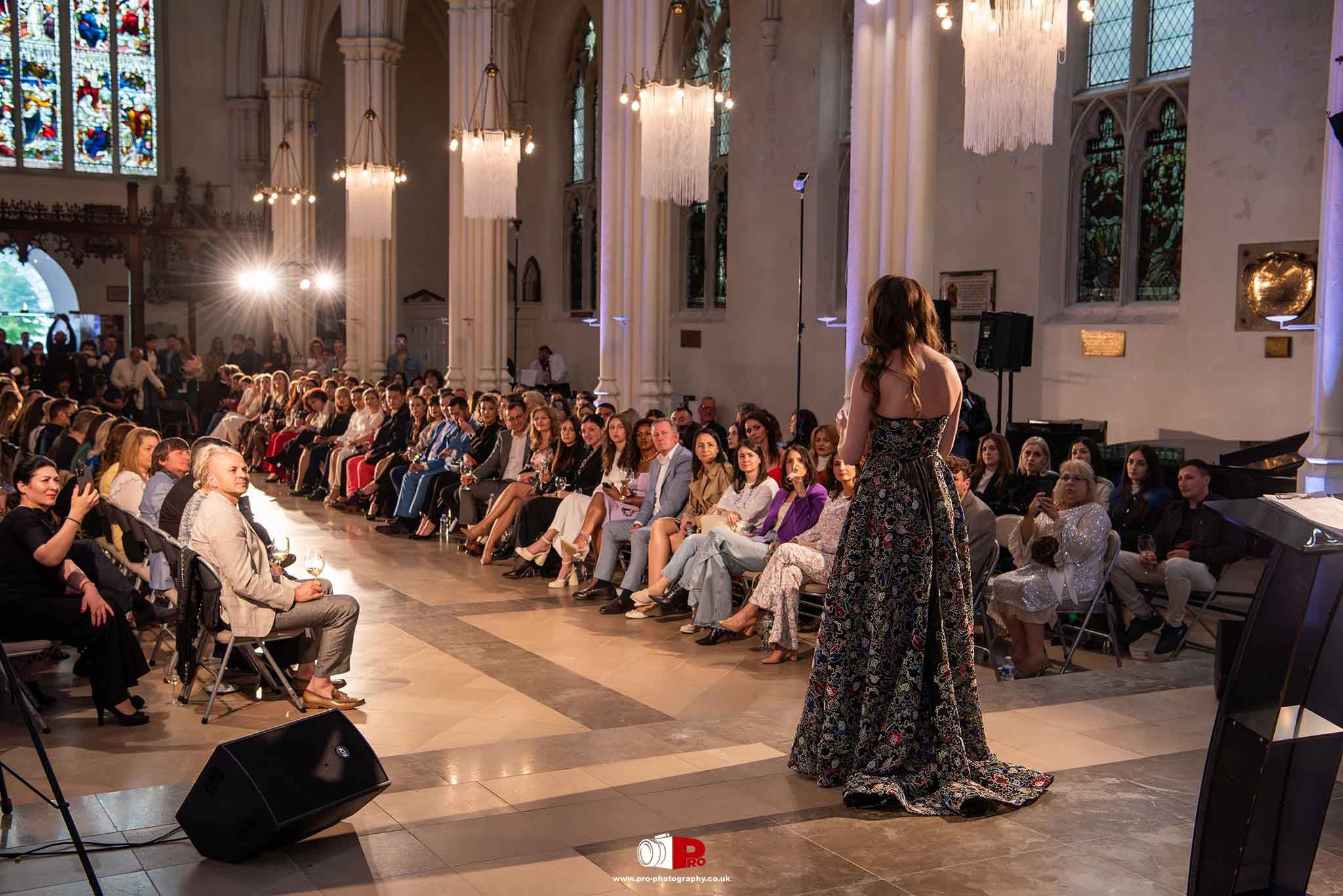 A broad view of the audience during a fashion show gala inside a beautifully lit venue with stained glass windows.