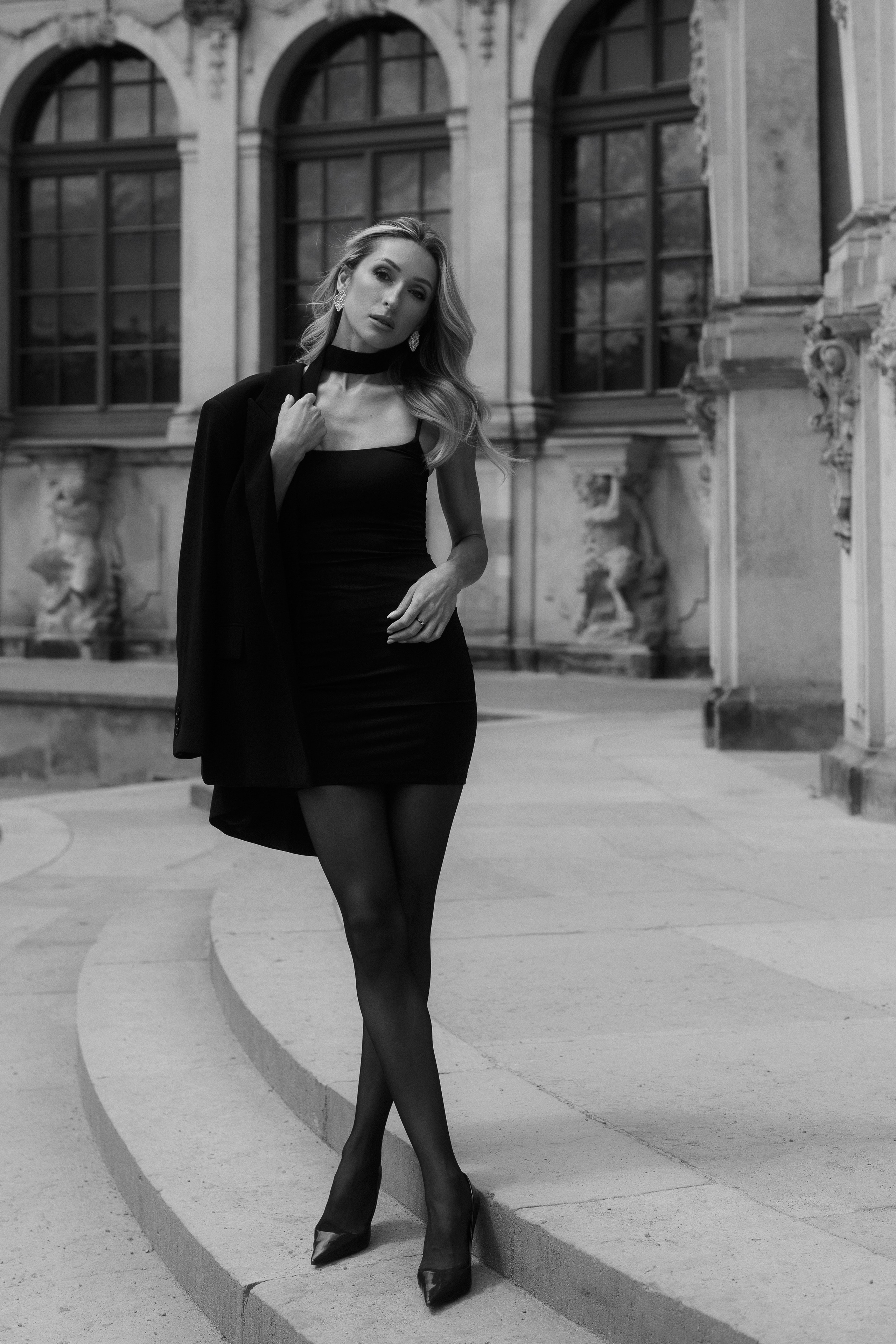 Elegant full-length black and white portrait of a woman in a black dress and blazer at a historic location in Dresden, Germany. Fine art and editorial portrait photography available in Dresden, Leipzig and Wurzen. Элегантный чёрно-белый портрет девушки в полный рост в чёрном платье и жакете на фоне исторической архитектуры Дрездена, Германия. Художественная и редакционная портретная фотография в Дрездене, Лейпциге и Вурцене.