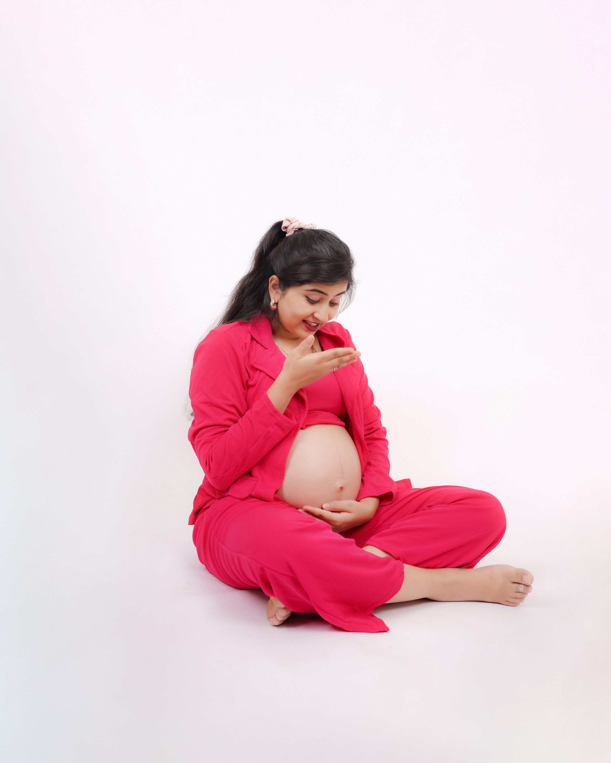 Maternity photoshoot in Bengaluru featuring a woman in a vibrant pink suit sitting cross-legged in an indoor studio, looking down at her baby bump.