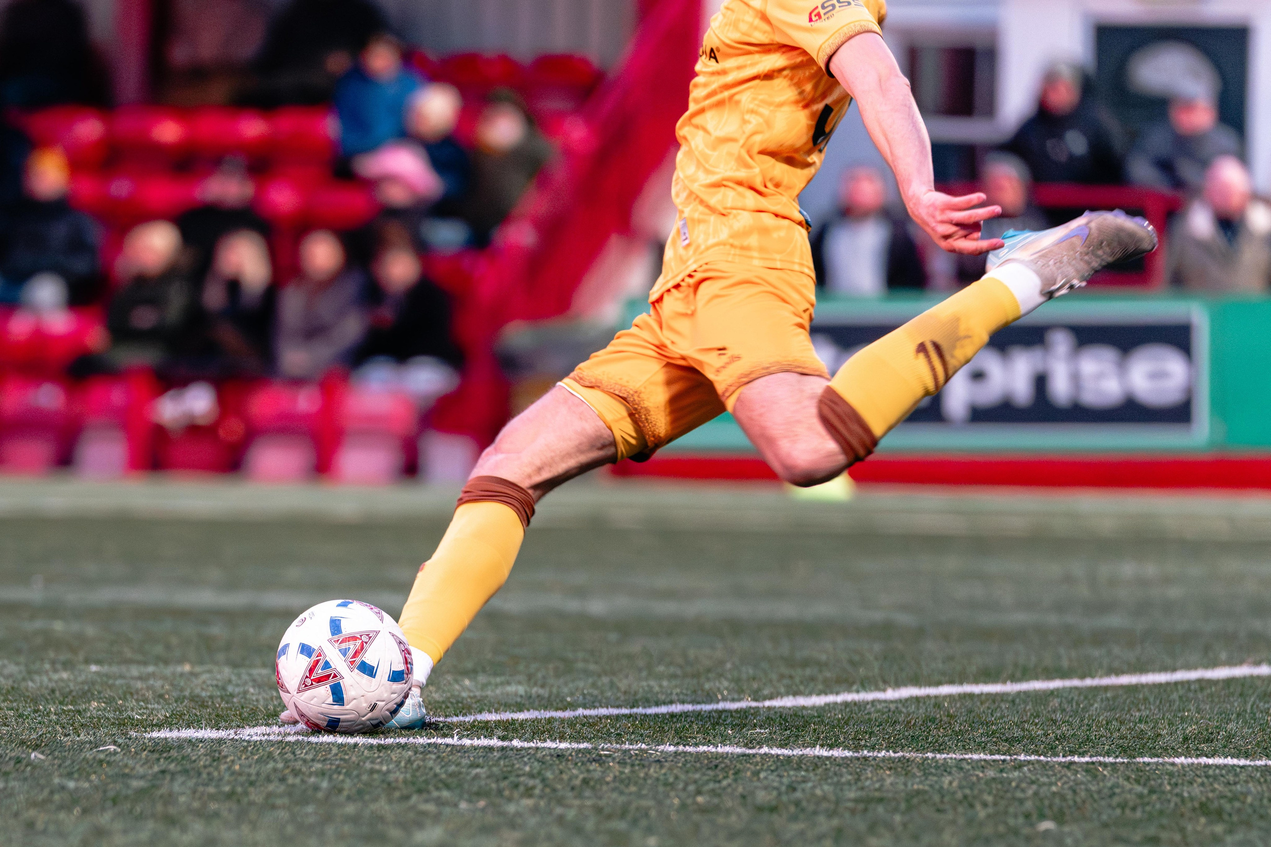 Sutton United player in yellow kit kicks the ball during open play on the pitch.