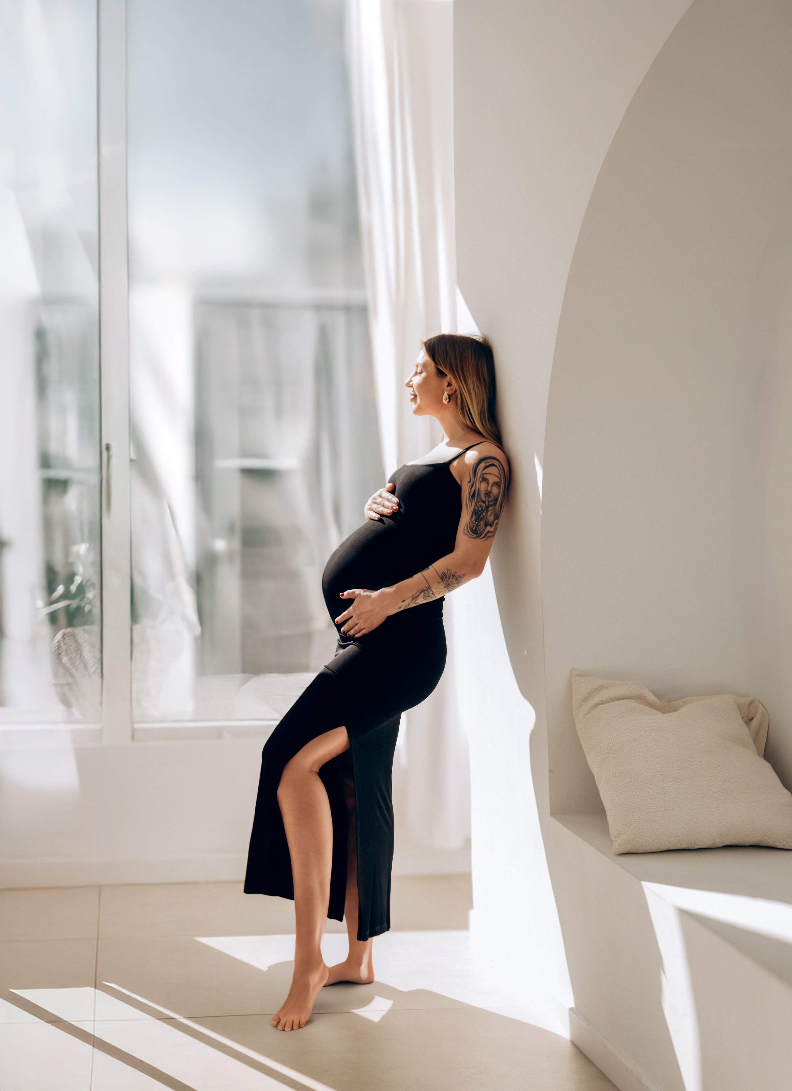 Retrato radiante de maternidad de una mujer embarazada con vestido negro, apoyada con elegancia contra una pared blanca iluminada por el sol en un estudio minimalista — capturando la belleza serena de la maternidad en Valencia, España.