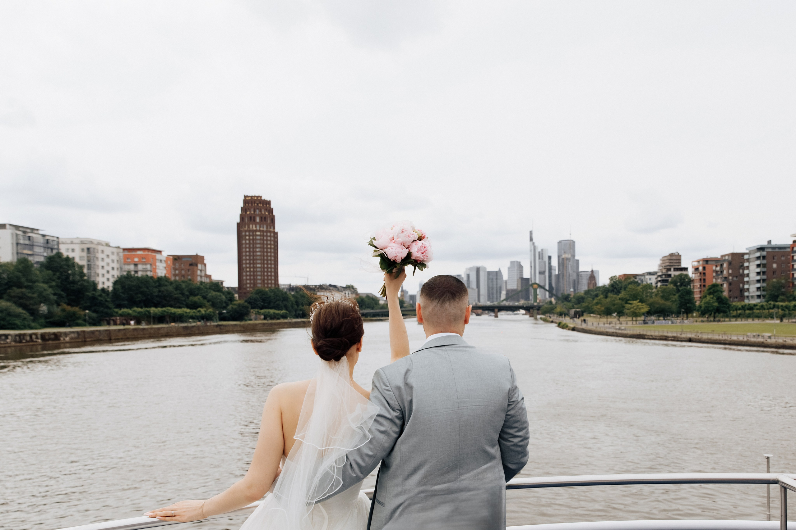 Der Bräutigam und die Braut schauen vom Schiff aus auf Frankfurt, während die Braut mit ihrem Brautstrauß winkt. 