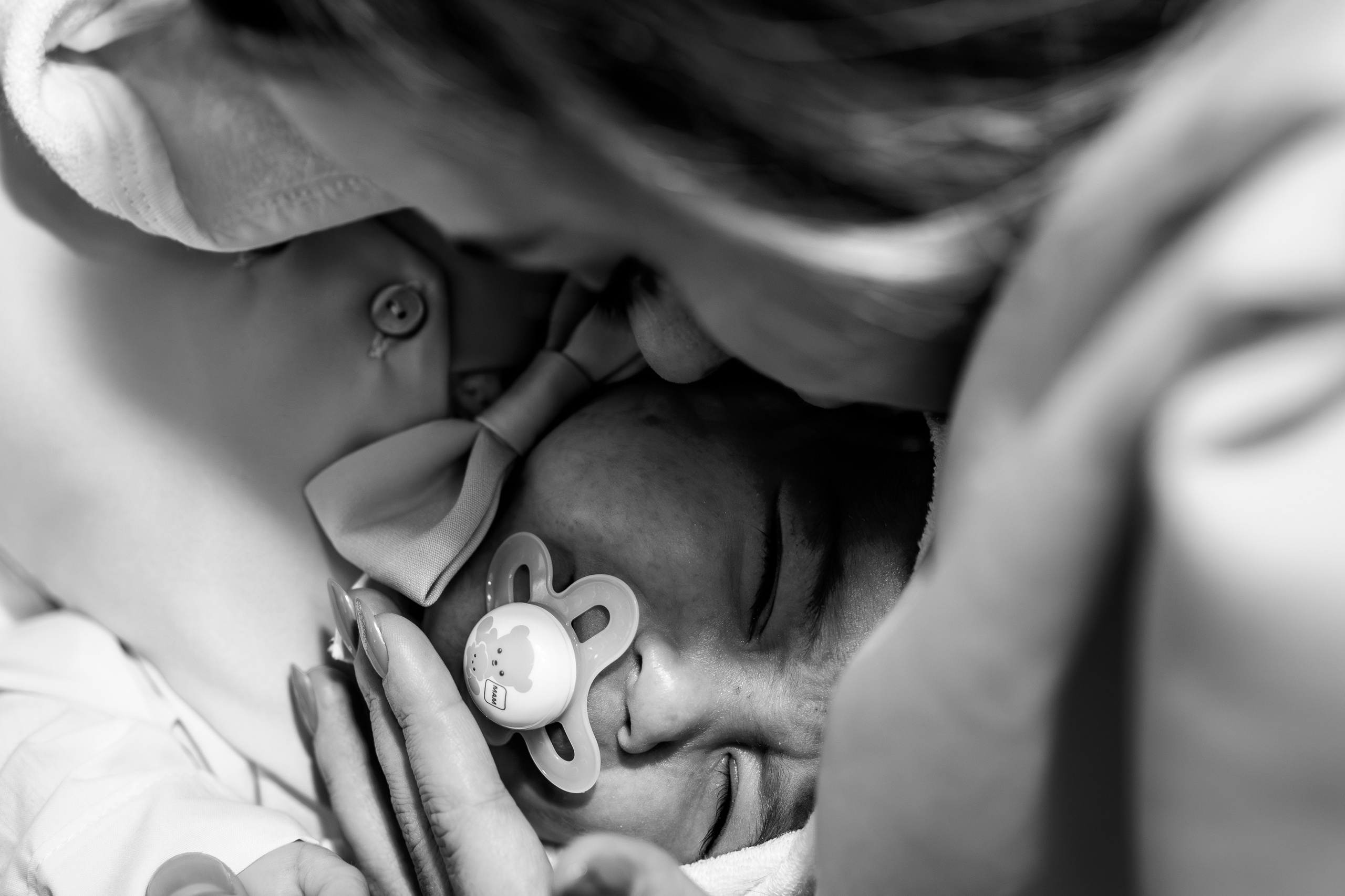 Fotografie alb-negru, close-up extrem de intim. Mama lui Arthur Mihail este foarte aproape de chipul pruncului, într-un cadru strâns care captează detaliul fiecărei gene, al respirației și al legăturii tăcute dintre ei, în pauza ceremoniei de botez.