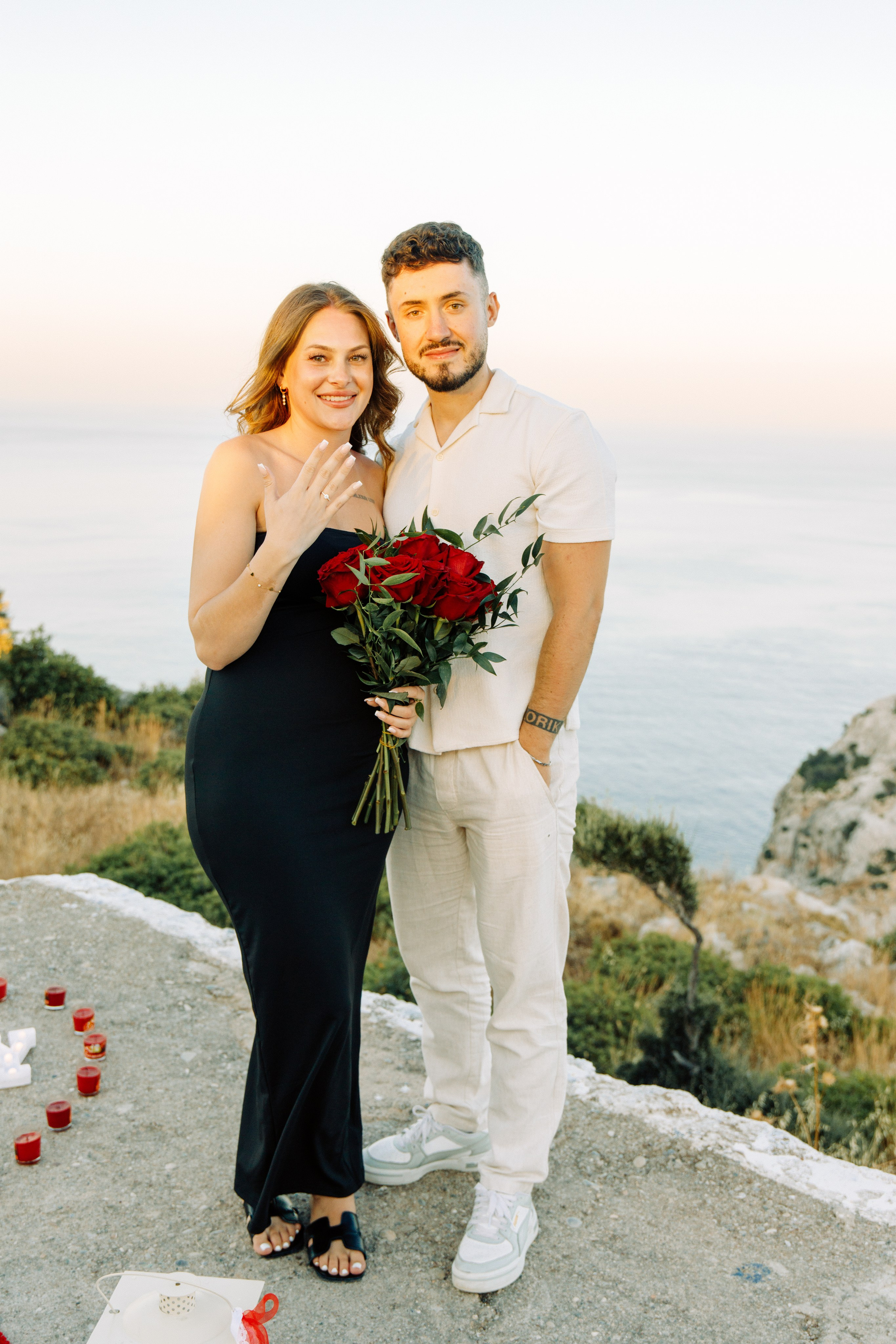 "Ein glückliches, frisch verlobtes Paar beim Fotoshooting auf dem Gipfel des Profitis Ilias. Sie stehen eng umschlungen auf einem Felsplateau, lachen in die Kamera und zeigen stolz den Verlobungsring. Im Hintergrund erstreckt sich das beeindruckende Panorama der Bucht von Faliraki und das glitzernde Ägäische Meer."