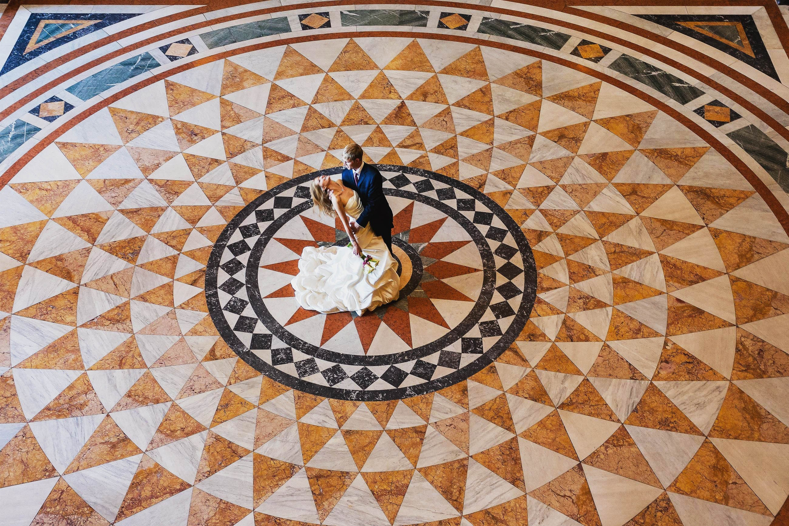 An American groom dips his beautiful blonde-haired bride in the center of a mosaic at the National Museum in Prague.