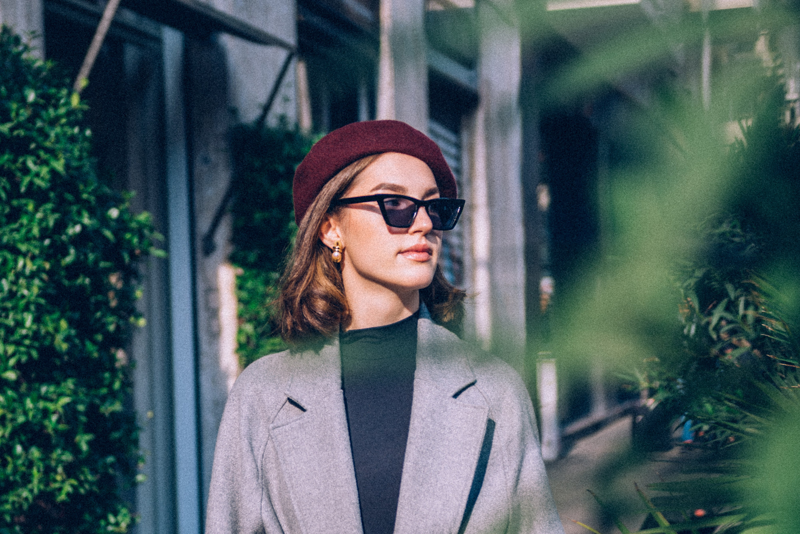 Urban portrait of a fashionable woman framed by greenery, dressed in a gray coat and accessorized with sunglasses and a beret. Emotional storytelling photographer in Milan