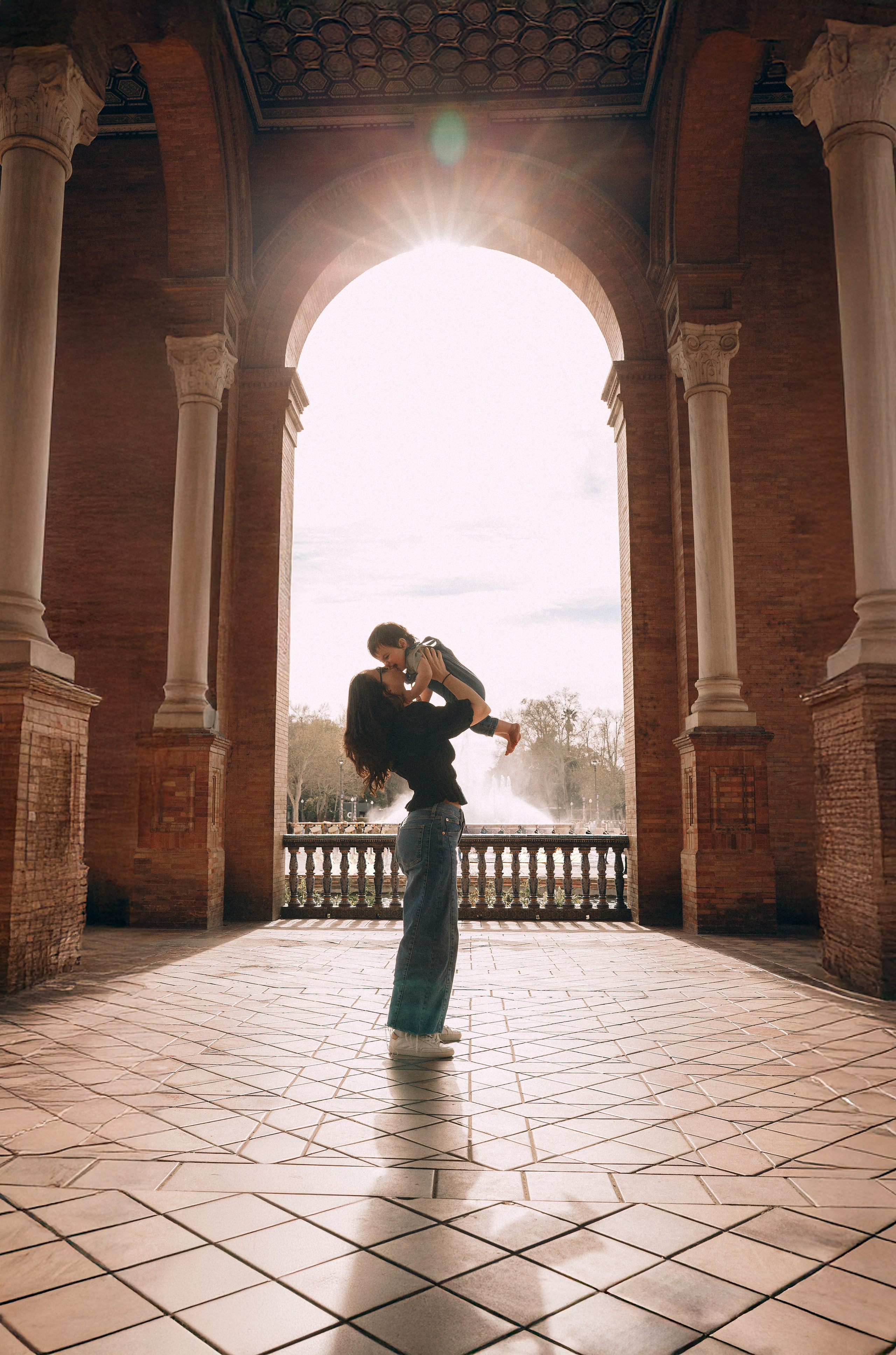 Momento familiar entrañable capturado en Sevilla, España — madre levantando a su hijo bajo los grandes arcos de la Plaza de España, con la luz del sol atravesando la arquitectura para crear un recuerdo atemporal y alegre.