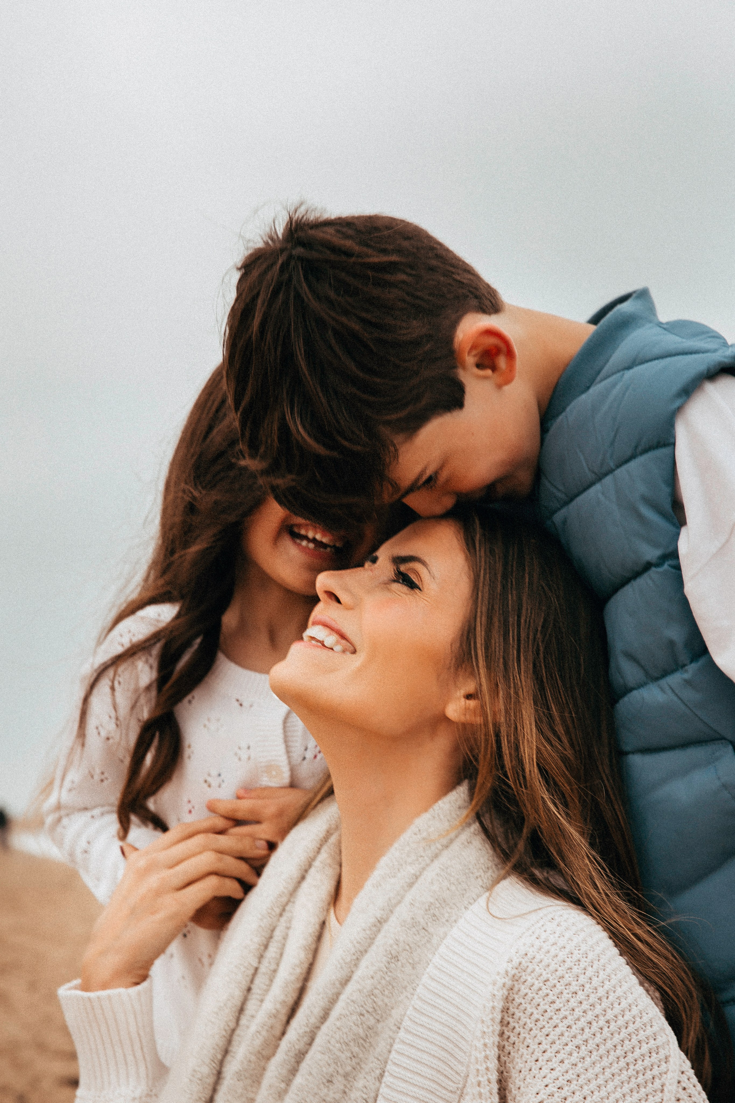 Momento familiar espontáneo en una playa ventosa de Valencia, España — madre sonriendo cálidamente mientras sus hijos la abrazan con amor y alegría. Una escena llena de conexión auténtica.