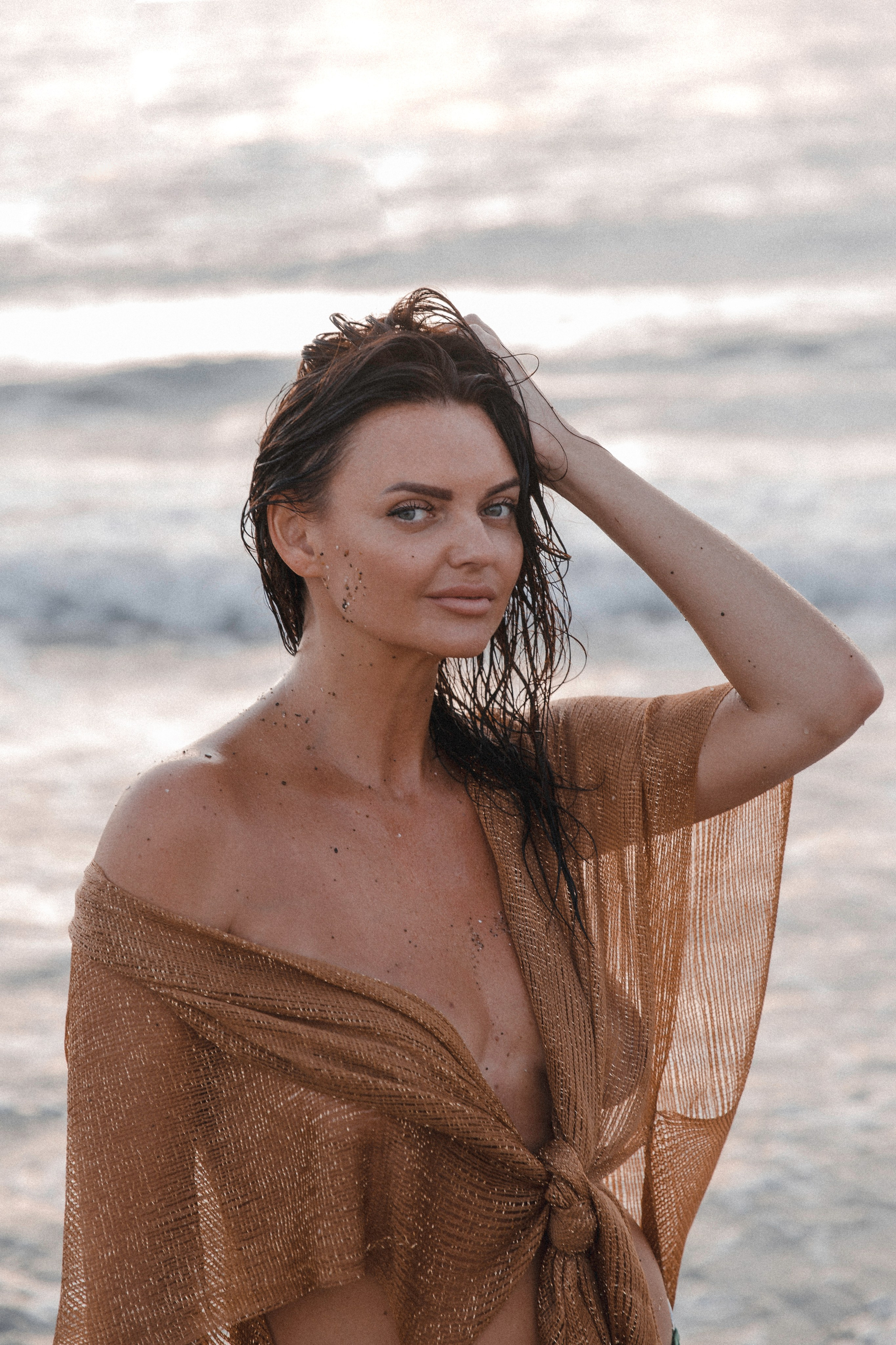 Гламурная женщина с мокрыми волосами и песком на коже, в золотистом прозрачном пляжном накидке, позирующая у моря на закате в Валенсии, Испания