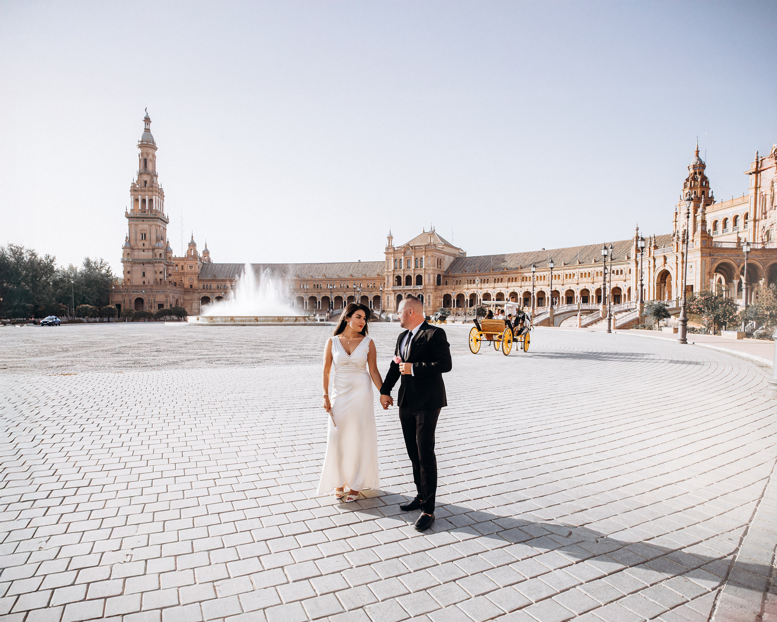 Bride and groom walking across the open square of Plaza de España in Sevilla under bright Spanish sunlight. Joyful scene from their intimate destination wedding celebration in Andalusia.