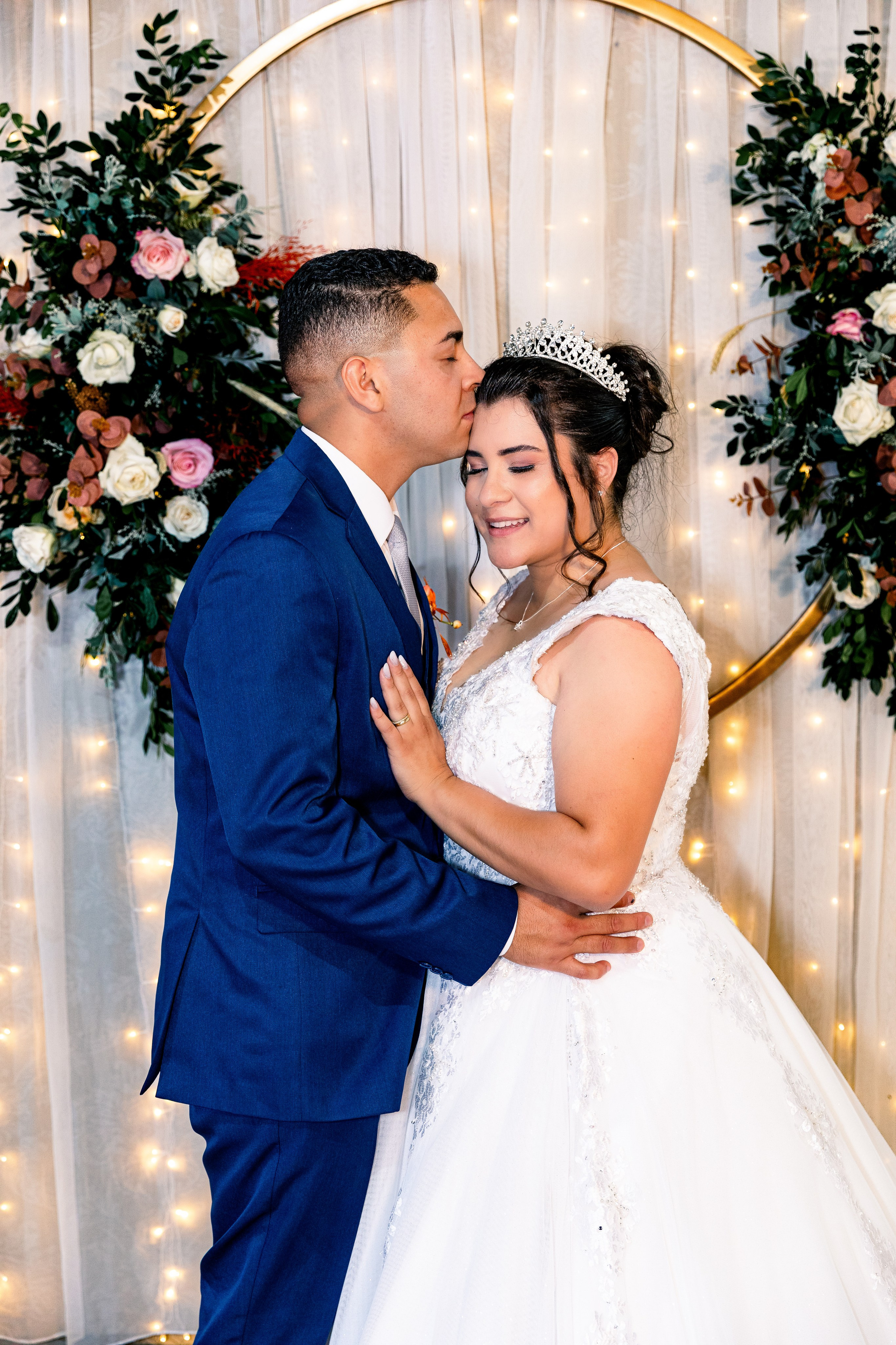 Noivo beijando a testa da noiva em frente a um painel decorado com flores e luzes, cena íntima e simbólica.