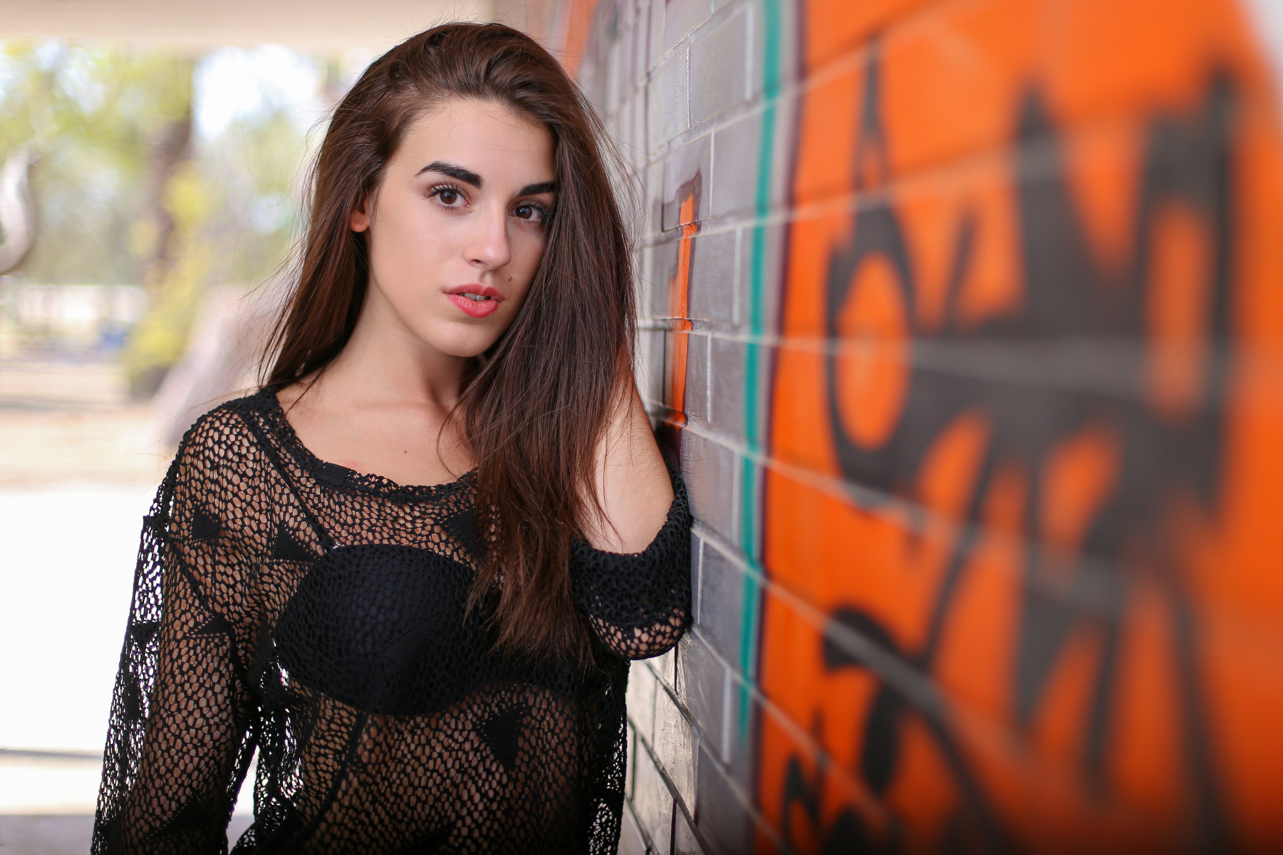 Urban fashion portrait of a young woman in a black sheer top posing by colorful graffiti wall.